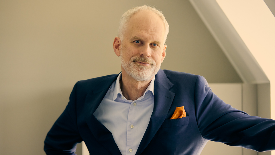 Porträttfoto av en äldre man med vit skägg, klädd i mörkblå kavaj och ljusblå skjorta