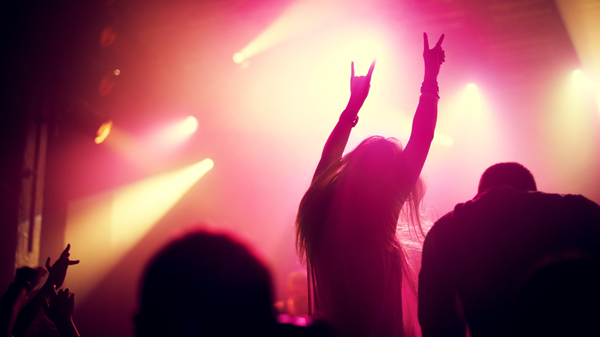 Silhuetter av människor på en konsert med upphöjda armar mot rosa och varmt scenljus.
