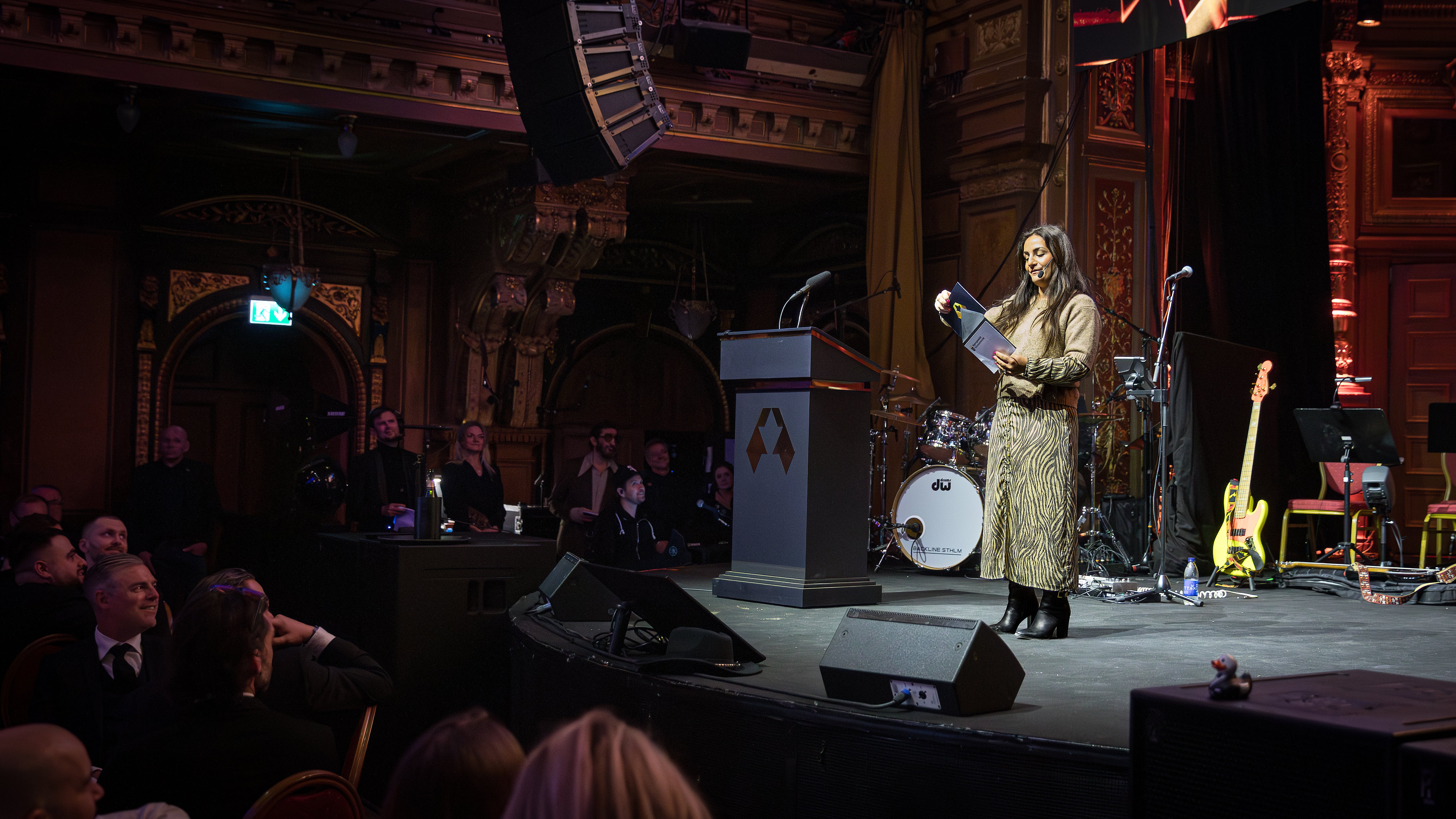 Kvinnlig artist på scen i guldklädd klänning talar vid mikrofon framför en orkester i en prydlig teatersalong med publik i förgrunden.