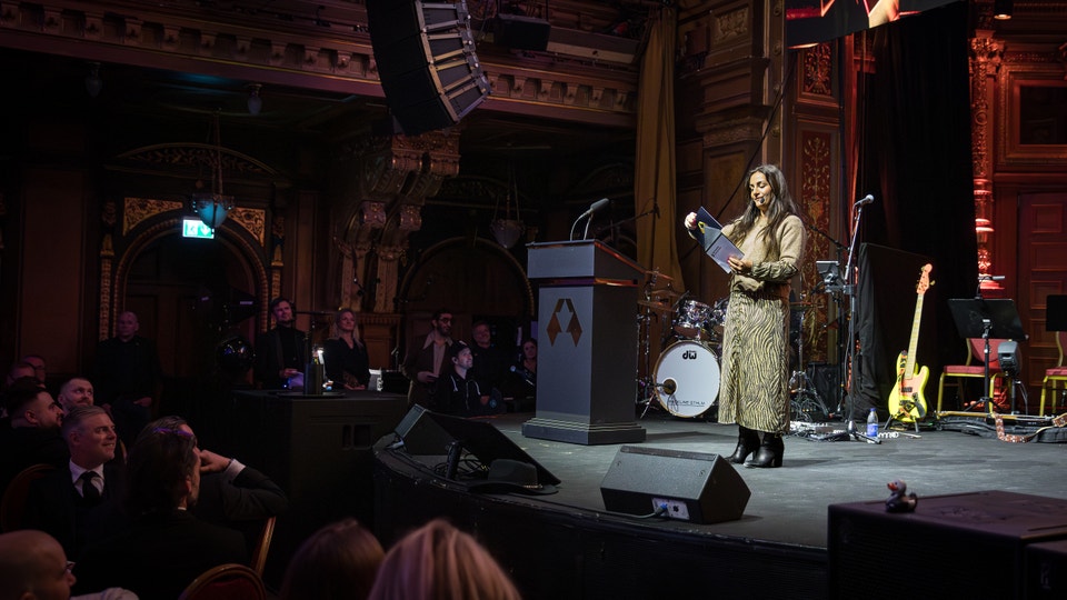Kvinnlig artist på scen i guldklädd klänning talar vid mikrofon framför en orkester i en prydlig teatersalong med publik i förgrunden.