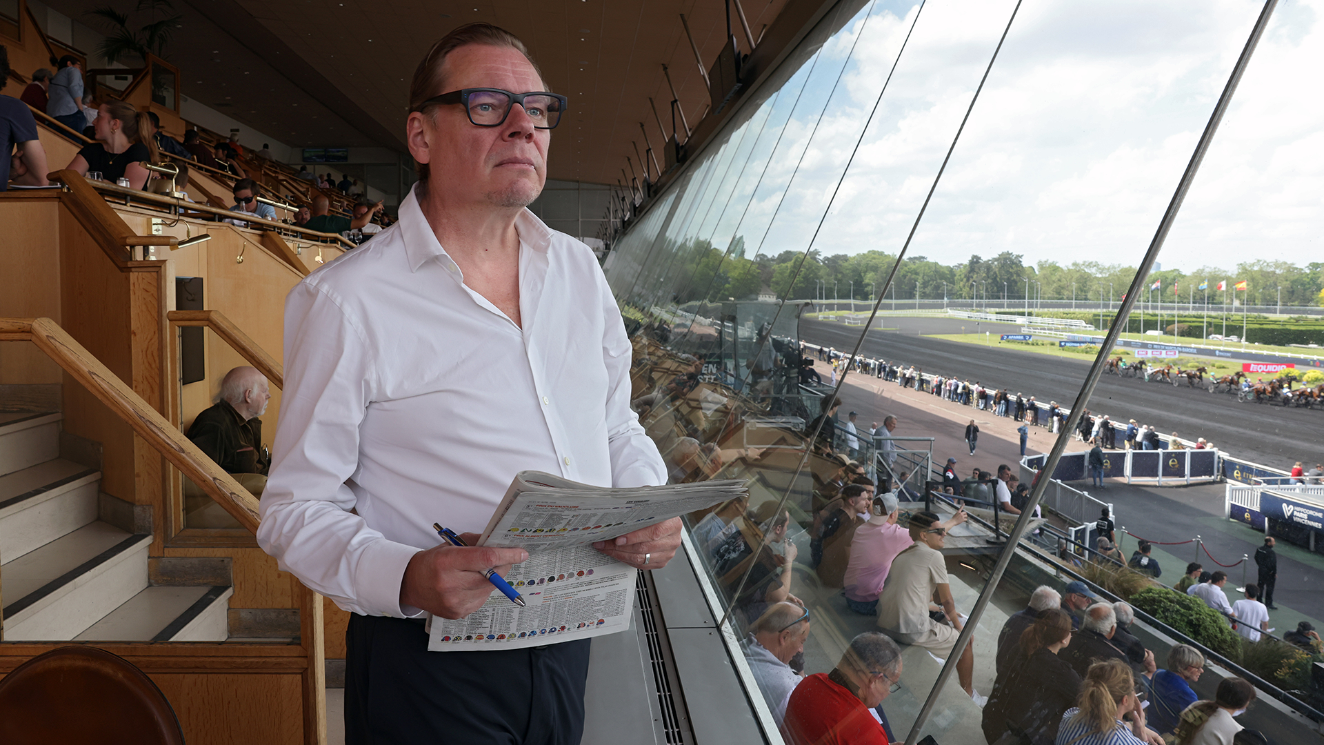 Man i vit skjorta och glasögon står i en tribuon på en racerbana och håller i programblad. Genom fönstret syns racerbanan med bilar och publik.