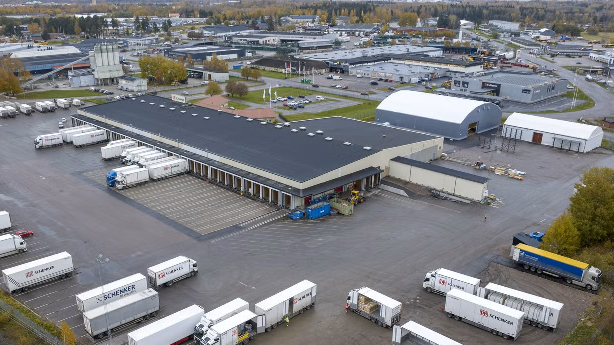 Flygfoto över ett logistikcentrum i Gävle med ett stort lagerbyggnad, lastningsplatser och parkerade lastbilar och släpvagnar på gården.