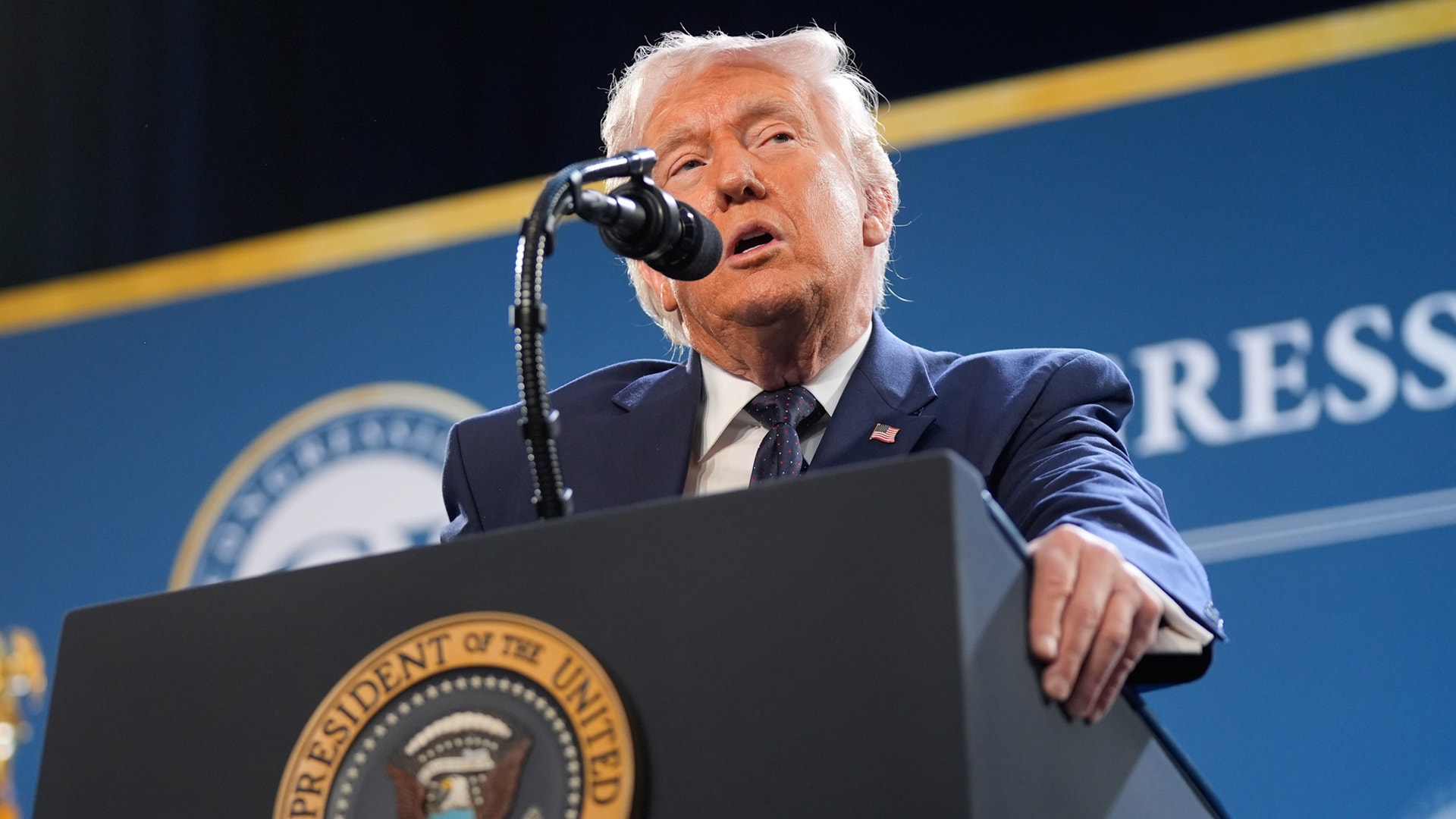 Man i mörk kostym talar vid ett podium med presidentens sigill framför blå bakgrund.