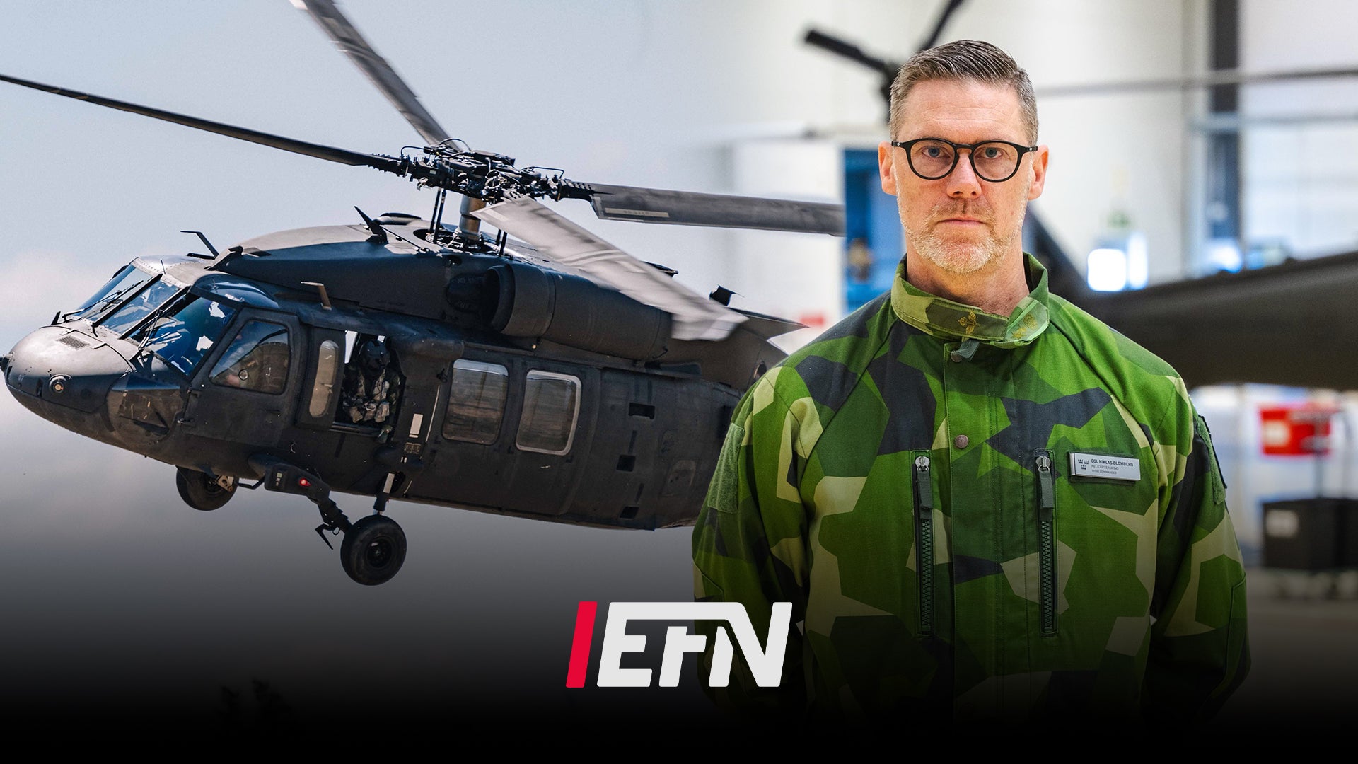 En militär helikopter och en soldat i grön kamouflageuniform står framför helikoptern i en hangar.