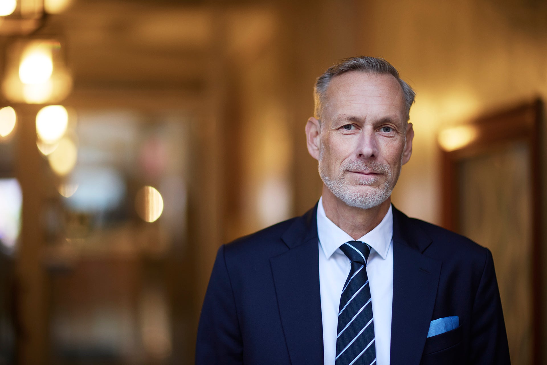 Porträtt av en man med grånande hår och kort skägg, klädd i mörkblå kostym, vit skjorta och randig slips. Han står i en varm, svagt upplyst inomhusmiljö.