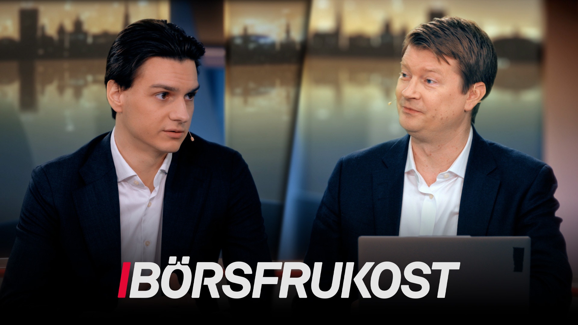 Två män i affärskläder diskuterar i en TV-studio med stadsutsikt i bakgrunden