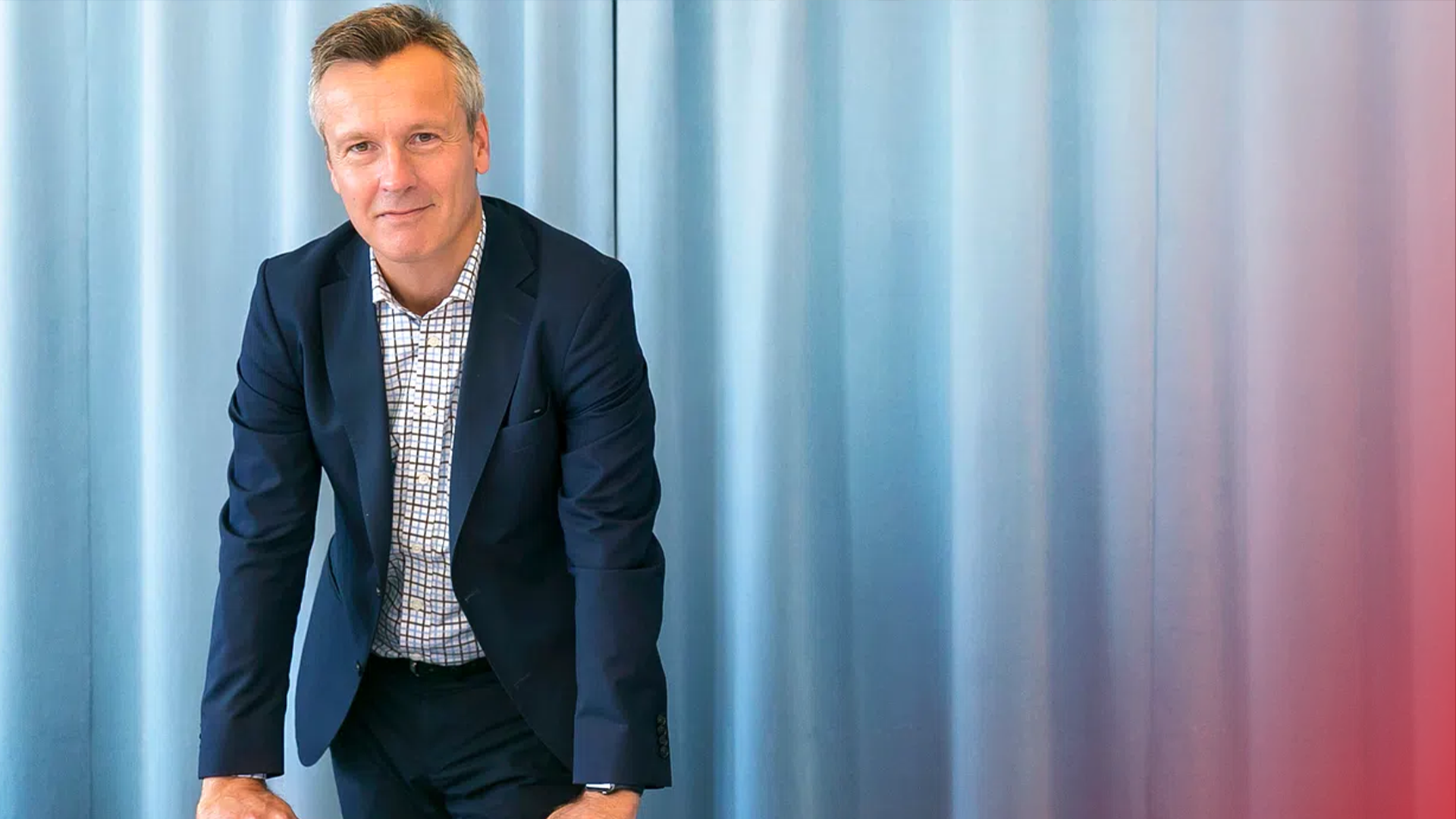 Porträtt av en man i mörkblå kostym och rutmönstrad skjorta mot en suddig bakgrund.