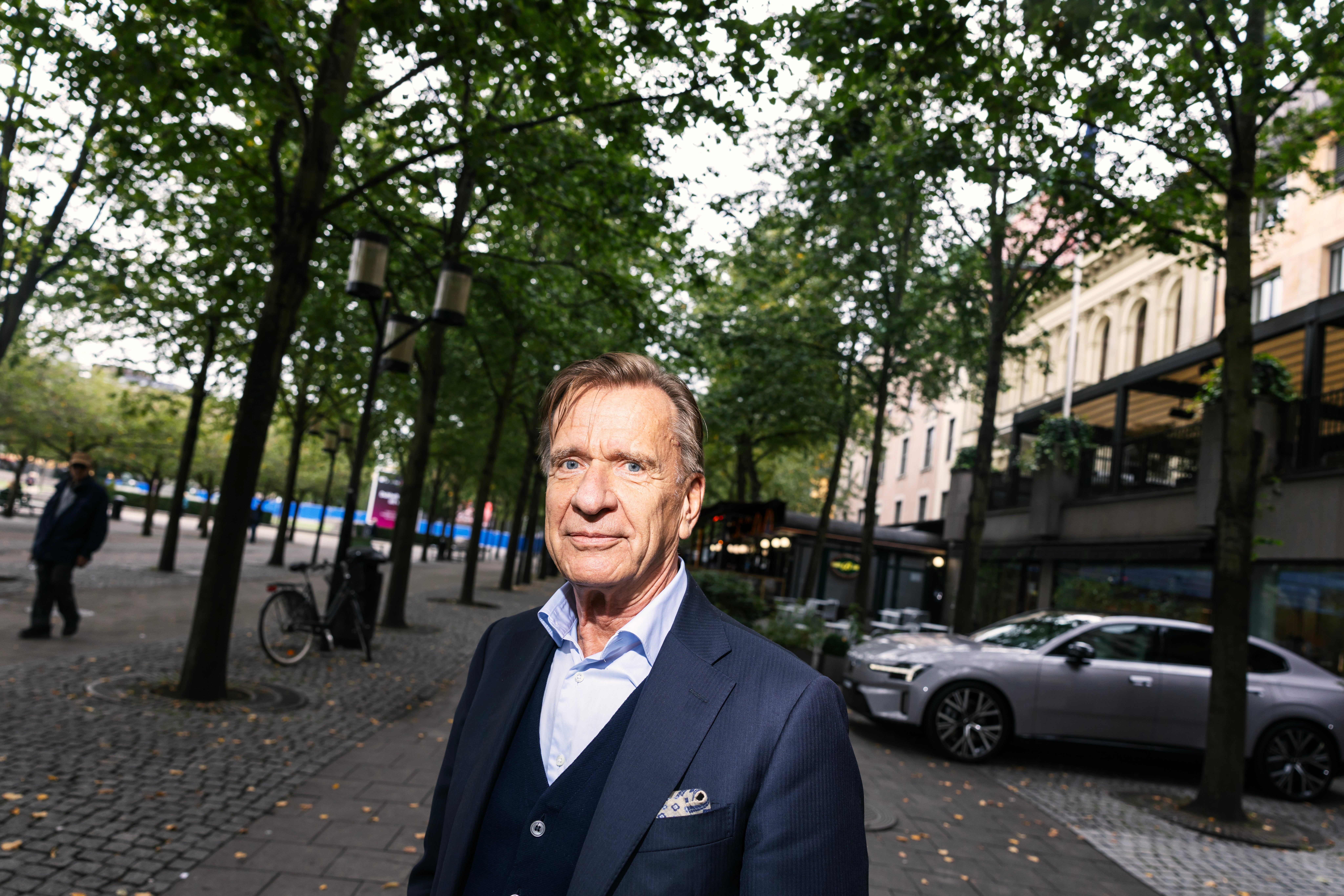 En äldre man i kostym står utomhus på en gata kantad av träd, med bilar och byggnader i bakgrunden.