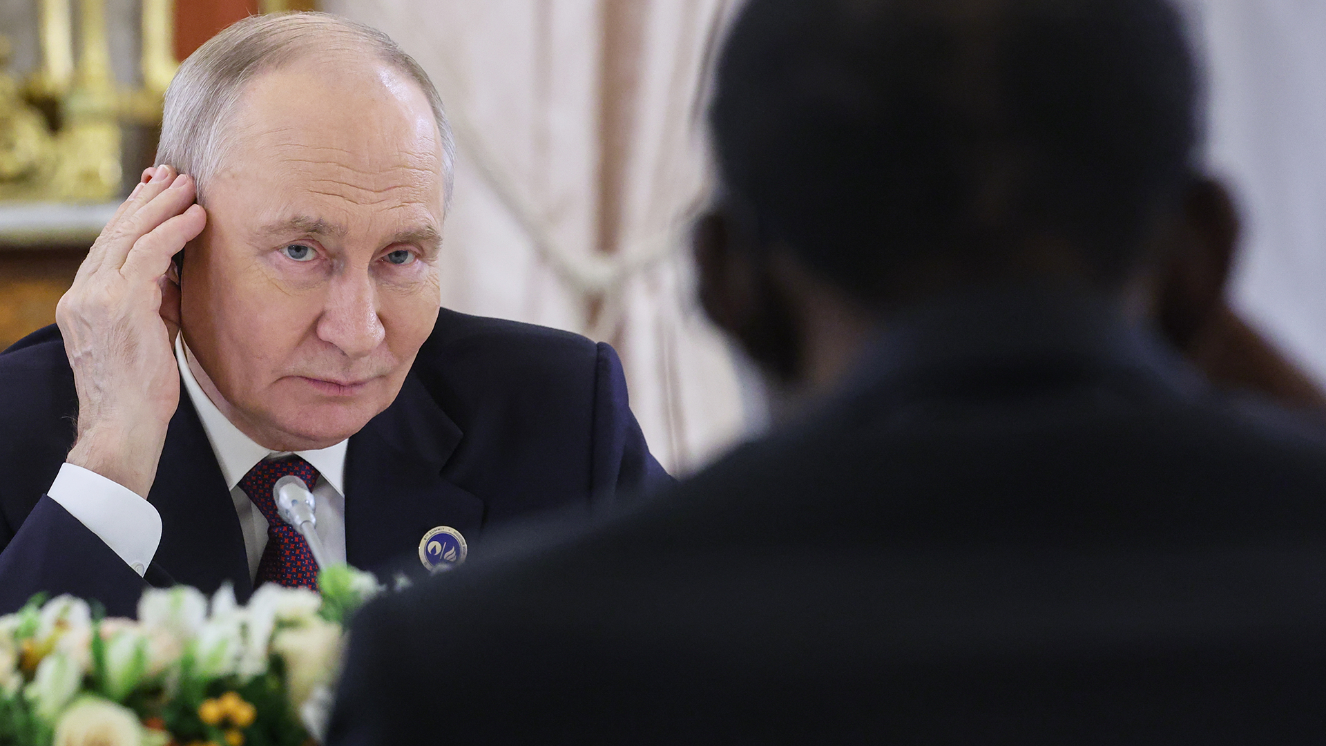 Vladimir Putin i mörk kostym lyssnar med handen vid örat under ett möte, medan en person i förgrunden syns bakifrån.
