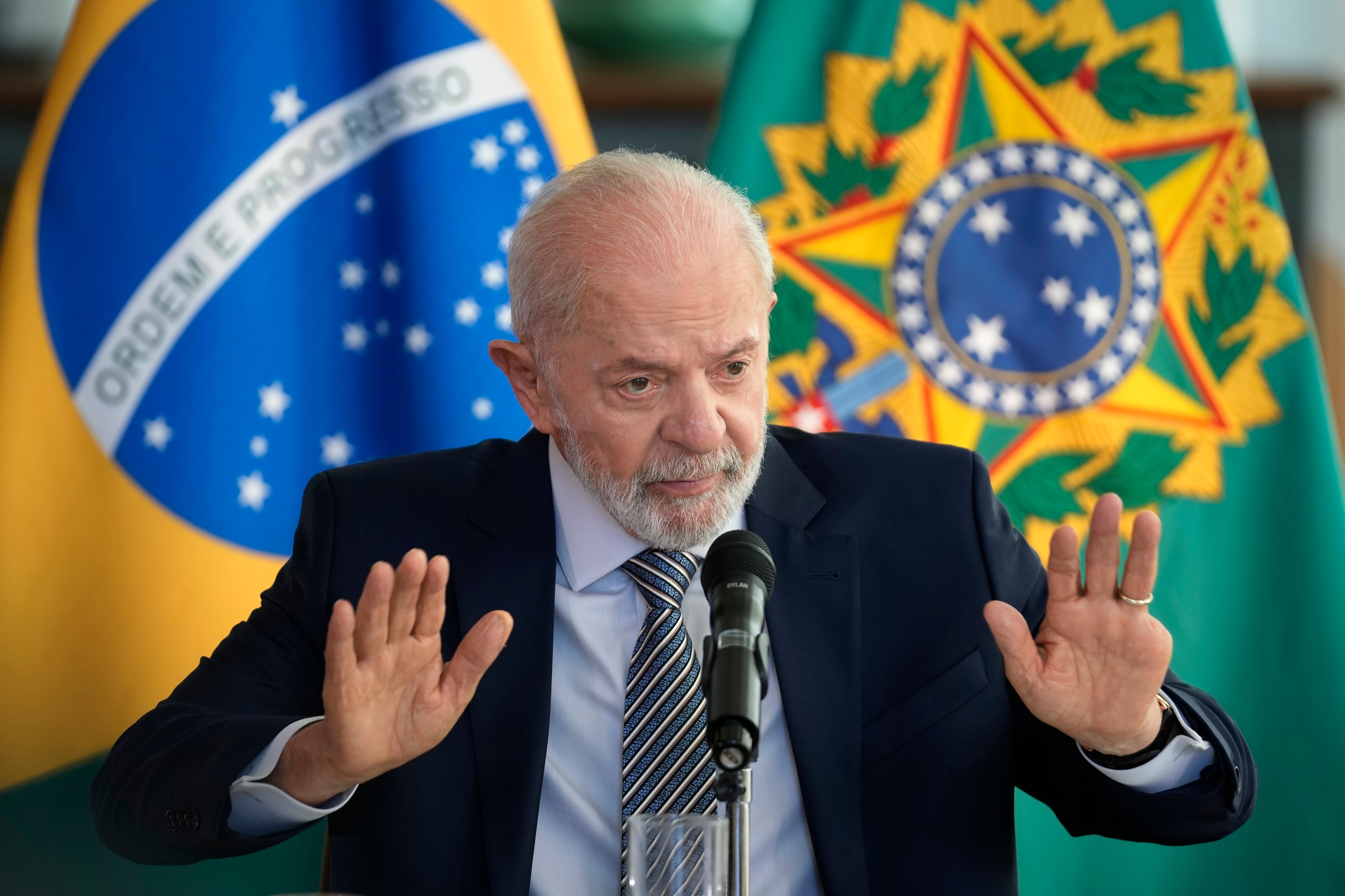 En man i kostym talar vid en mikrofon med gester, framför brasilianska flaggor.