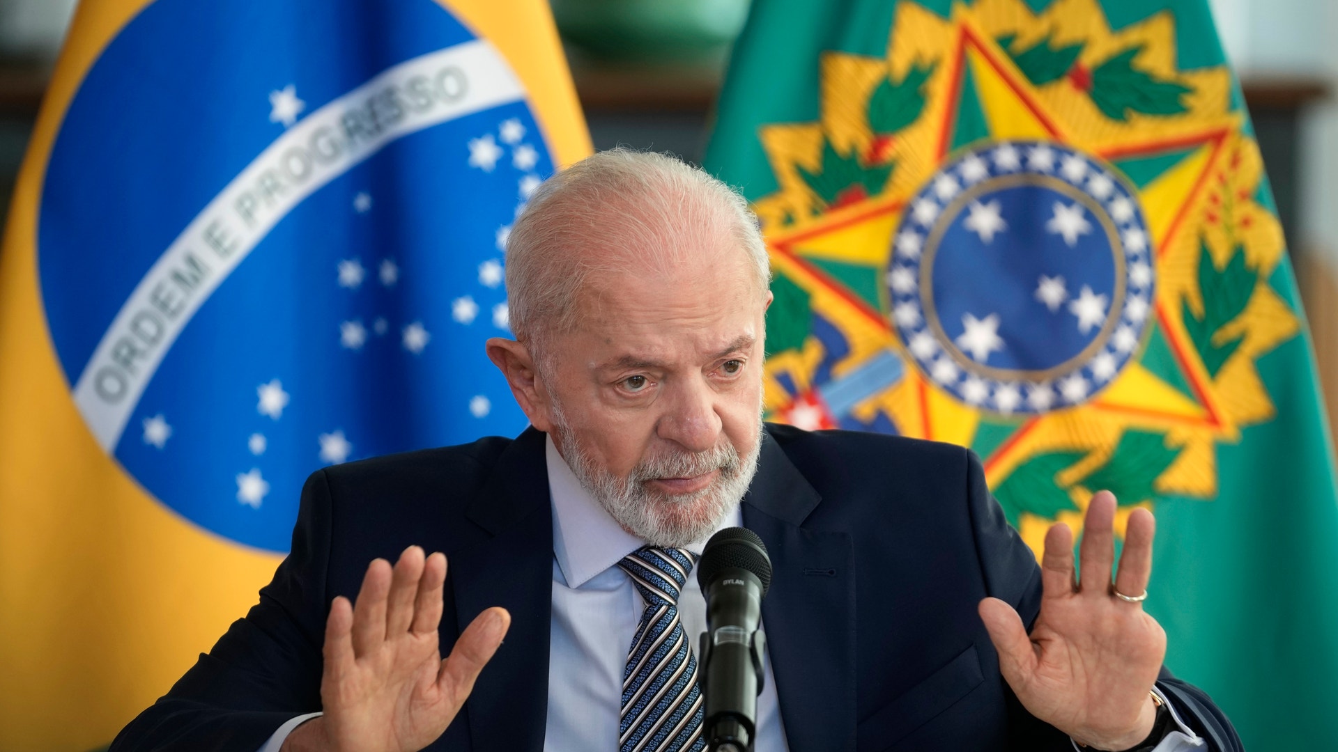 En man i kostym talar vid en mikrofon med gester, framför brasilianska flaggor.