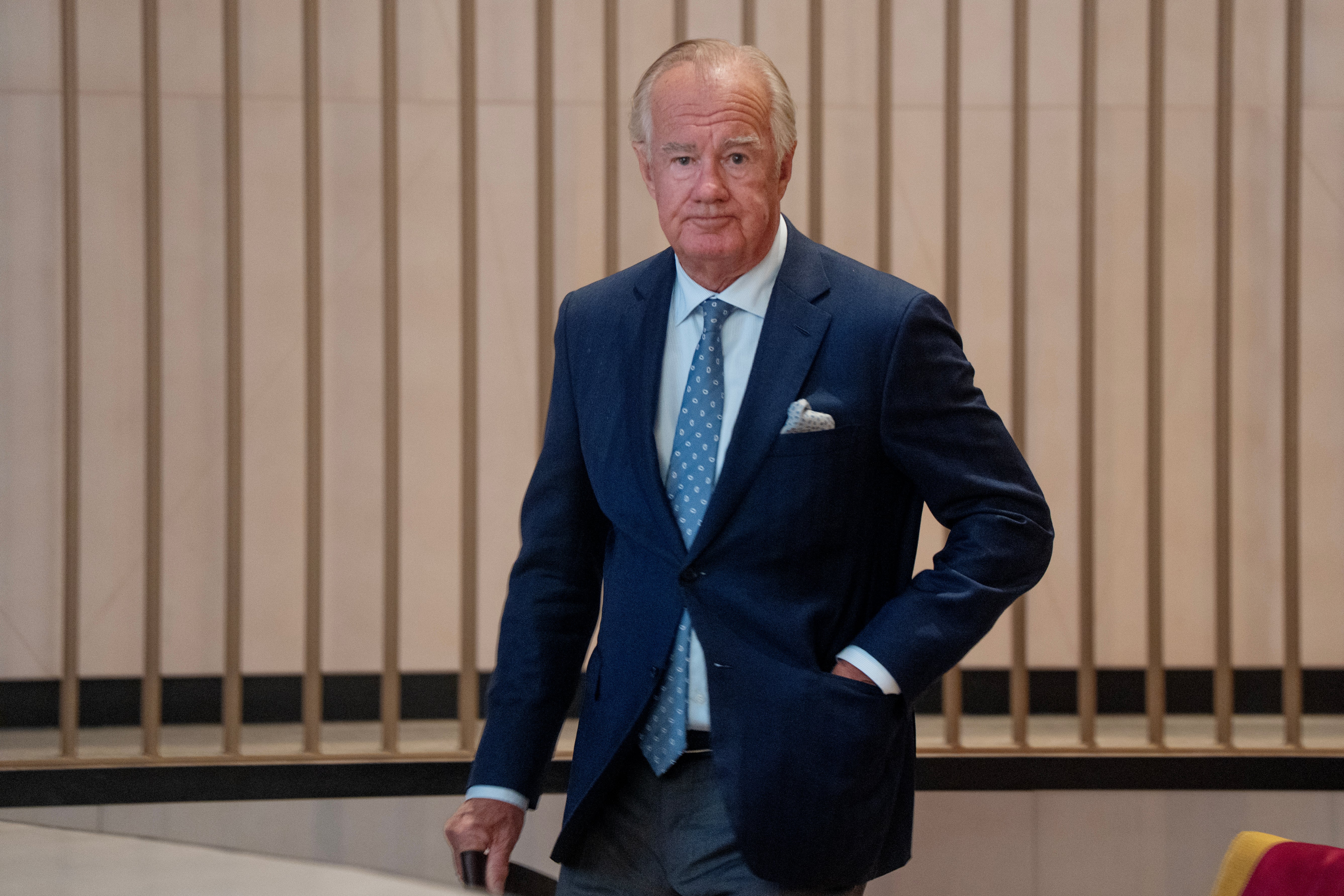 En äldre man i mörkblå kostym och blå slips står framför en vägg med träpaneler.