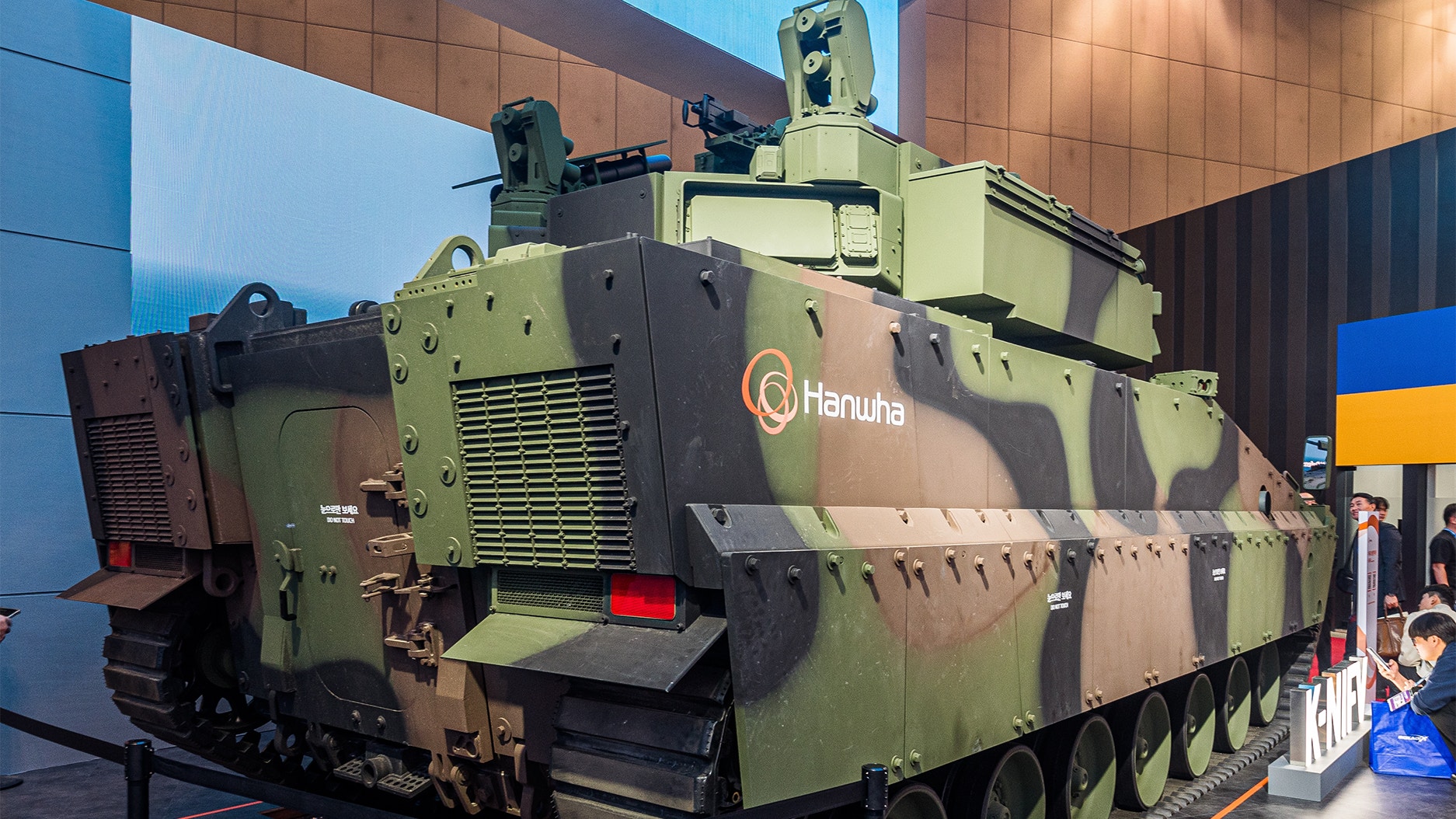 Militärt stridsfordon från Hanwha i kamouflage, utställt inomhus med människor i bakgrunden.