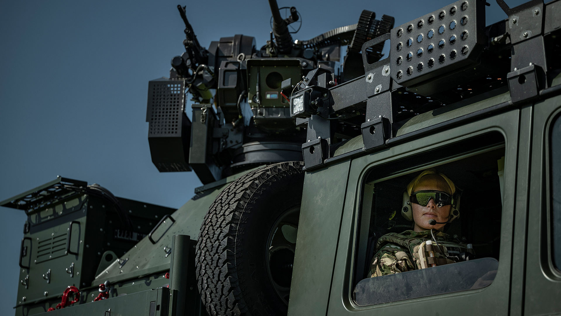 Soldat i militär utrustning sitter i ett militärt fordon och bär hjälm och skyddsglasögon. Fordonet är utrustat med vapen och militär utrustning på taket.