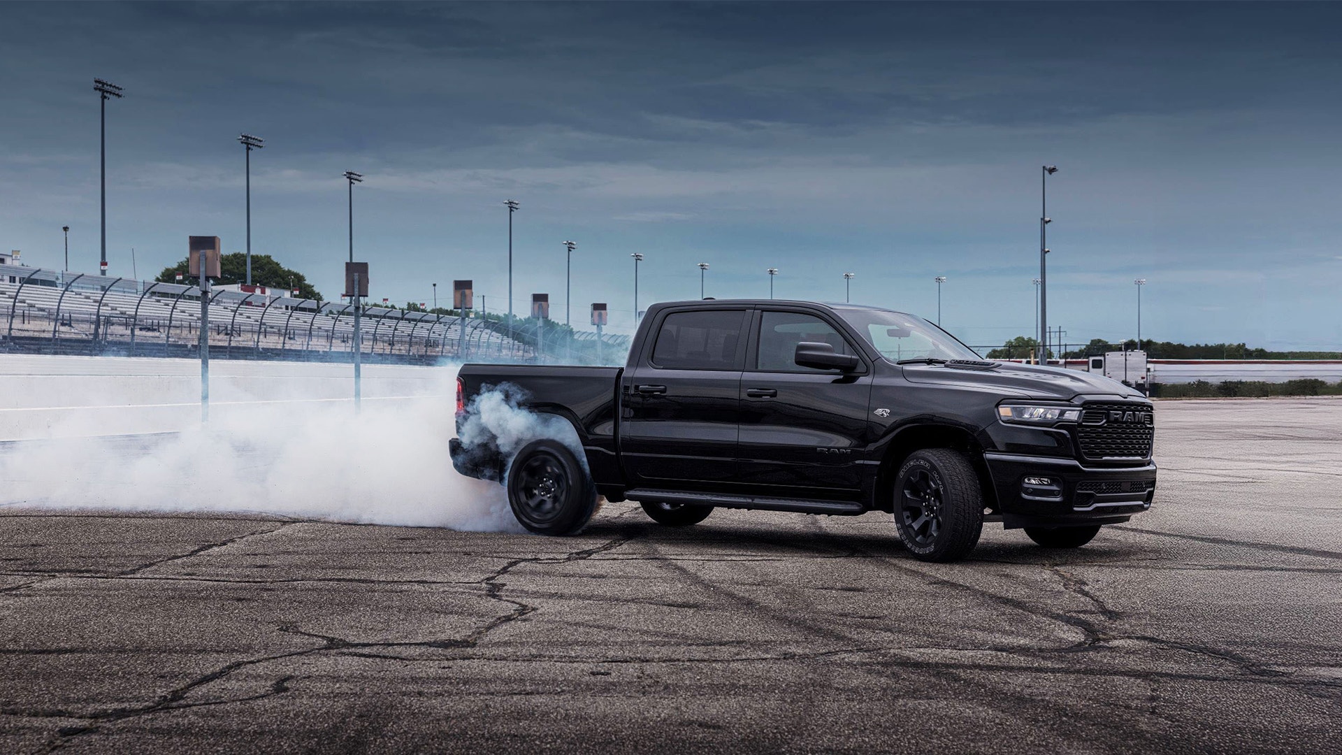 Svart pickup-truck som gör burnout med rök från däcken på en asfaltyta. Racerbana med belysning syns i bakgrunden.