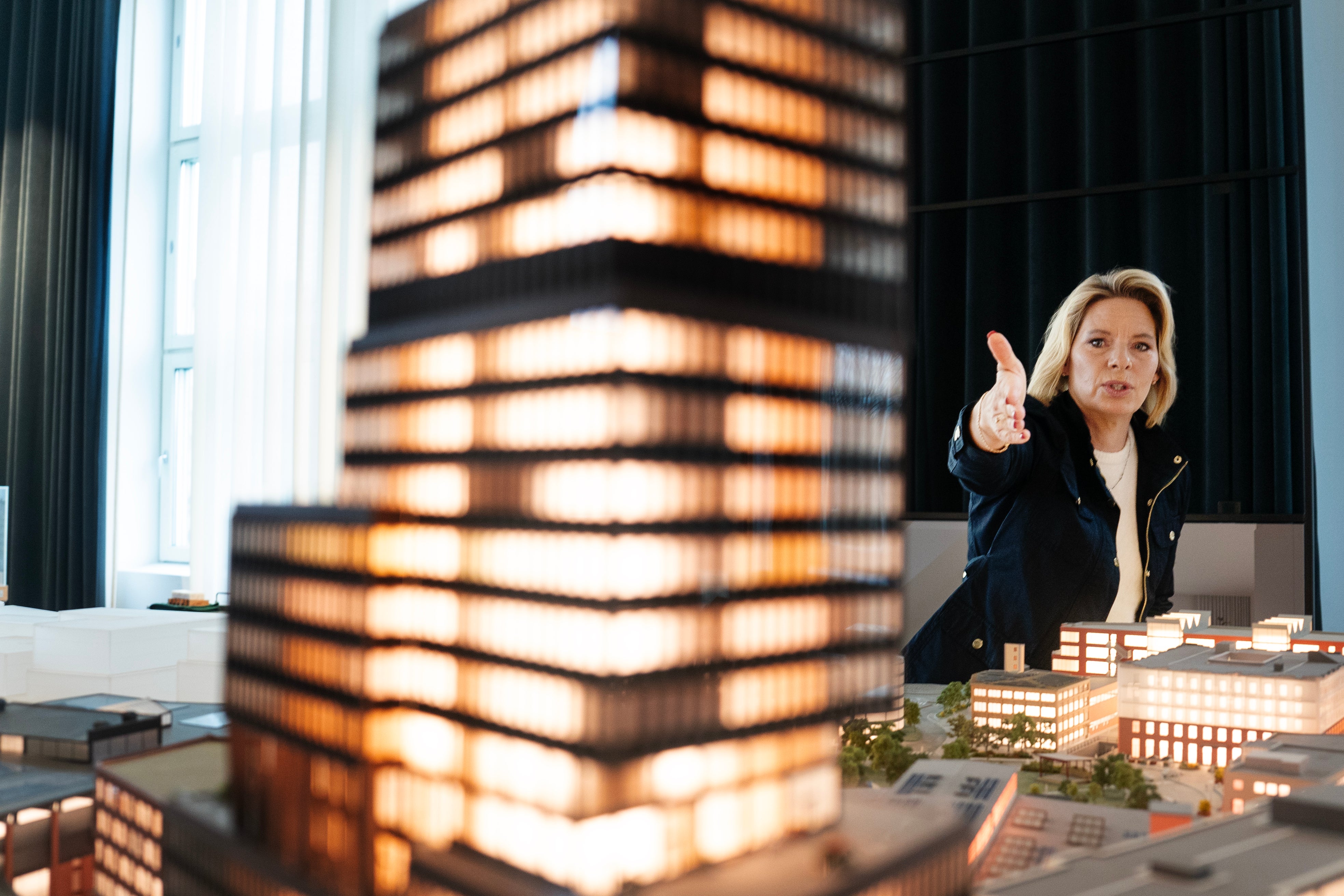 En kvinna presenterar arkitekturmodeller av byggnader på ett bord. Hon pekar och tittar upp medan hon diskuterar de belysade modellerna framför sig.