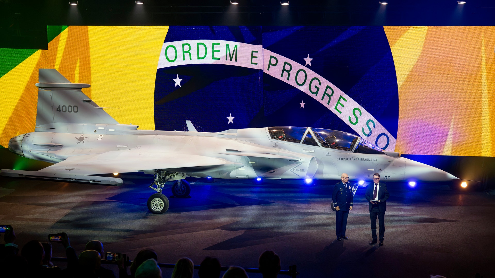 Ett modernt stridsflygplan presenteras på en ceremoni framför den brasilianska flaggan. Flygplanet är belyst med blå ljus och människor tittar på från förgrunden.