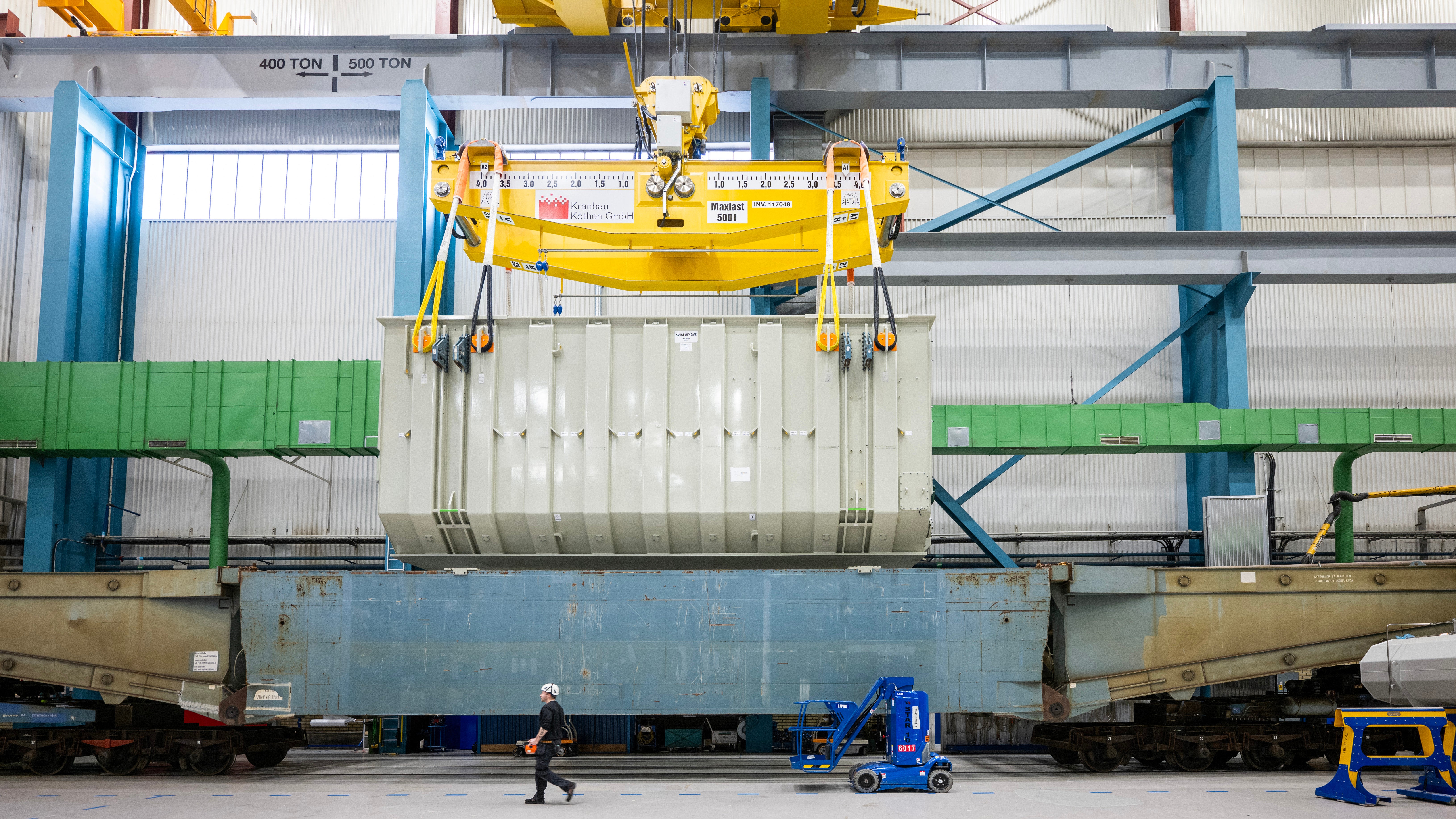En stor transformator lyfts med kran ner på en tågvagn inne i en industrihall.