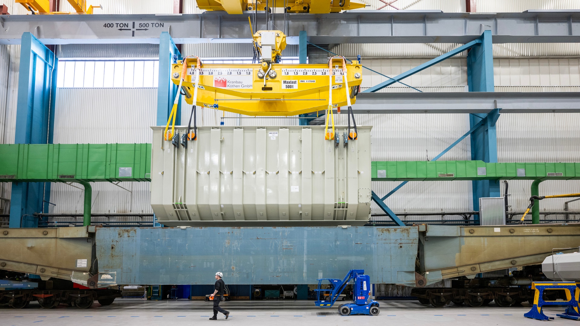 En stor transformator lyfts med kran ner på en tågvagn inne i en industrihall.
