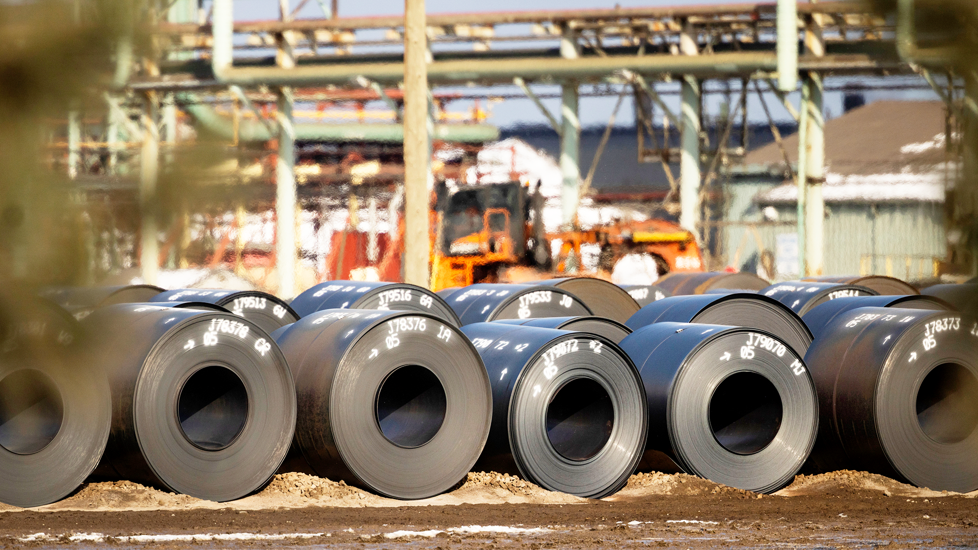 Rullar av stålspolar staplade på rad vid en industrianläggning med maskiner och byggnader i bakgrunden.