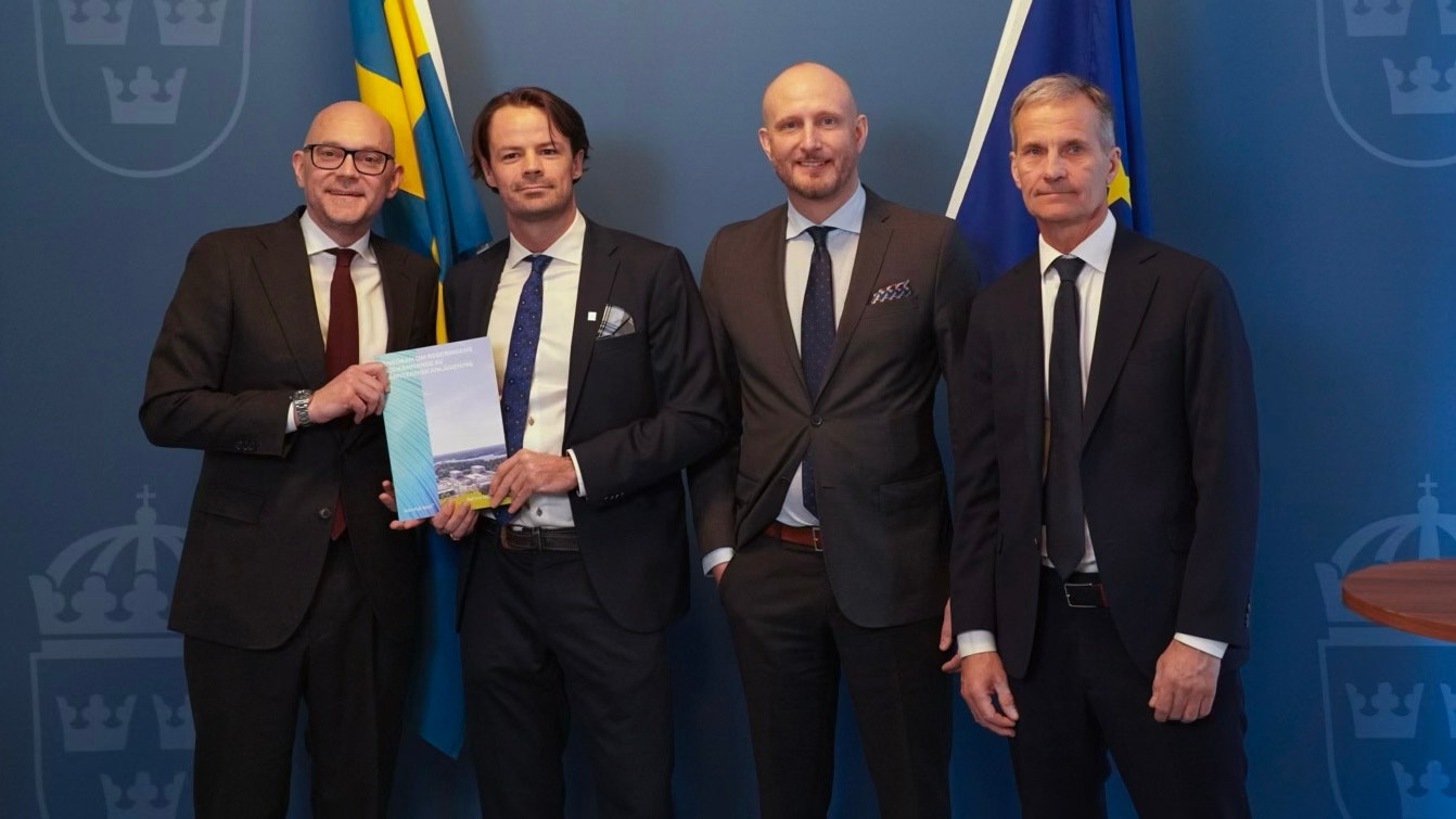 Fyra män i kostym står framför blå bakgrund med svenska flaggor. Två av dem håller ett certifikat eller diplom.