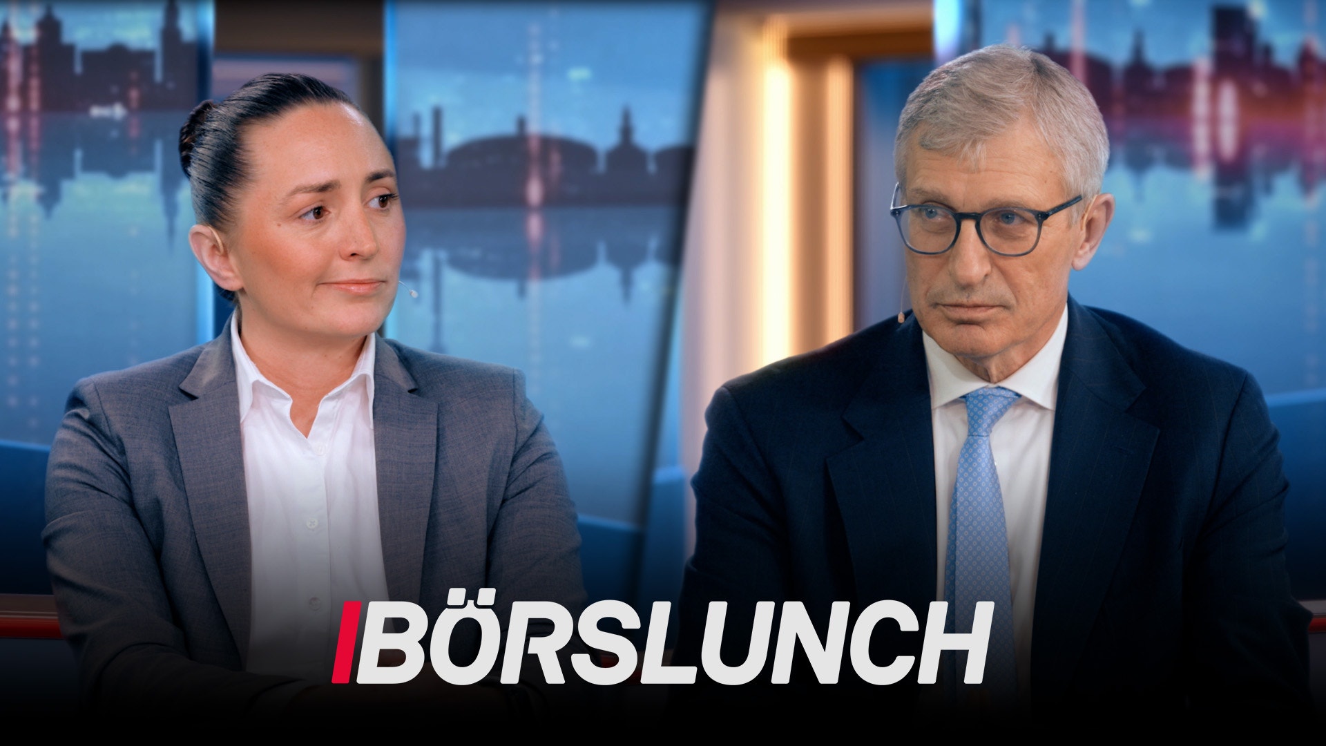Två personer sitter vid ett studiobord i TV-programmet Börslunch. I bakgrunden syns en stadsskyline. Personen till vänster är en kvinna i grå kavaj och vit skjorta, och personen till höger är en man i mörk kavaj och blå slips med glasögon.