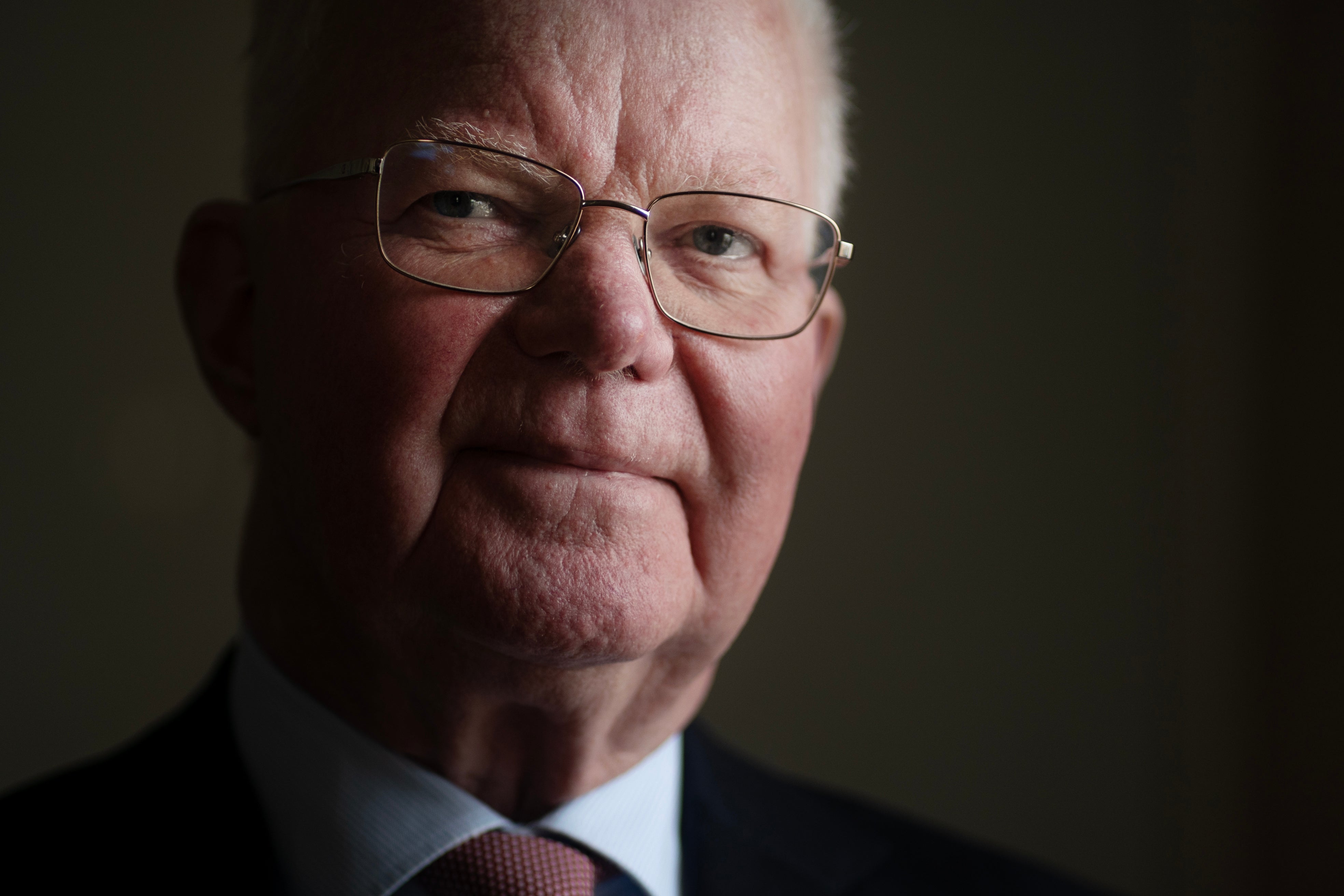 Porträtt av en äldre man med glasögon, iklädd mörk kostym och röd slips.