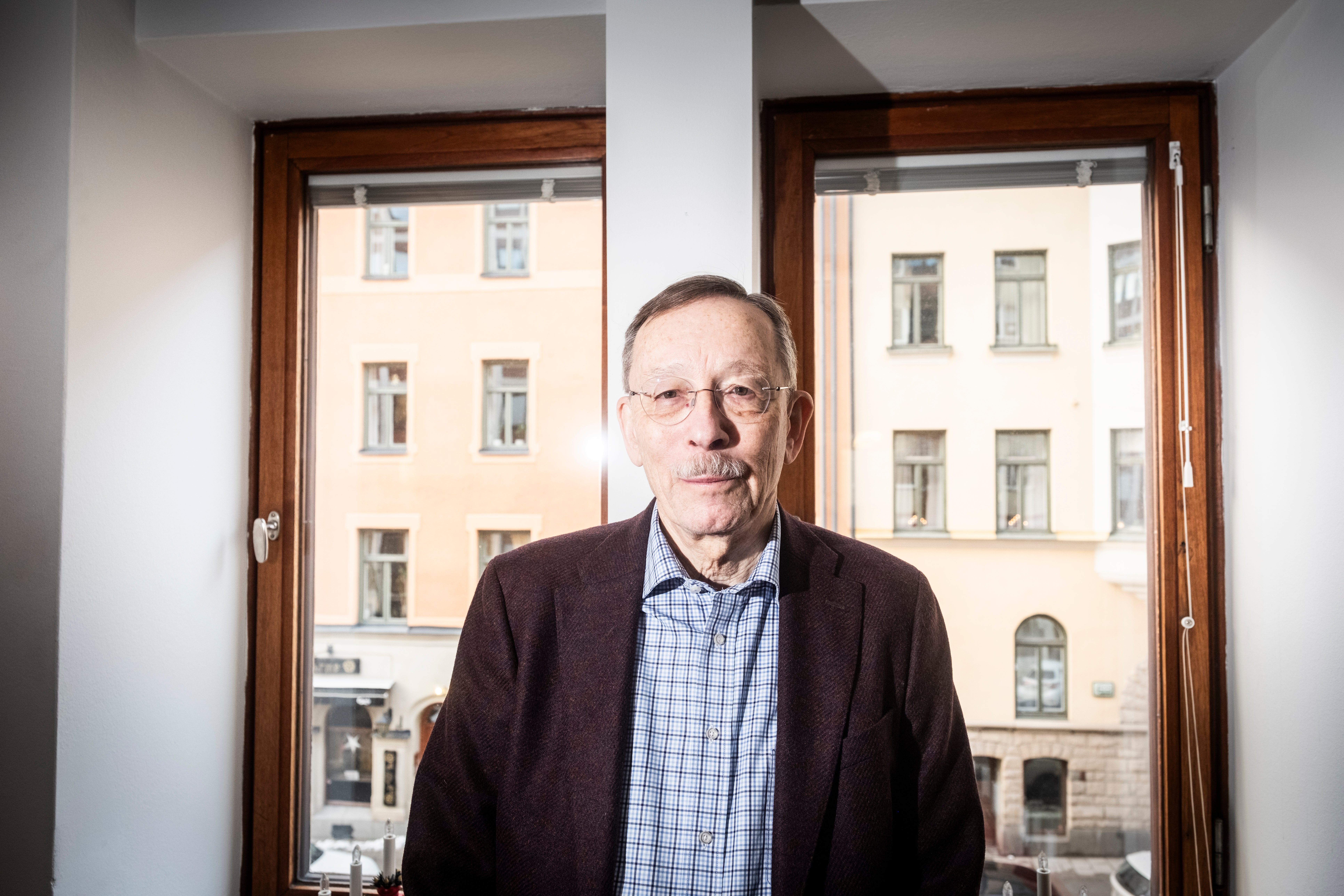 Porträtt av en äldre man med glasögon och mörkbrun kavaj som står framför ett fönster med utsikt över en stadsbyggnad.