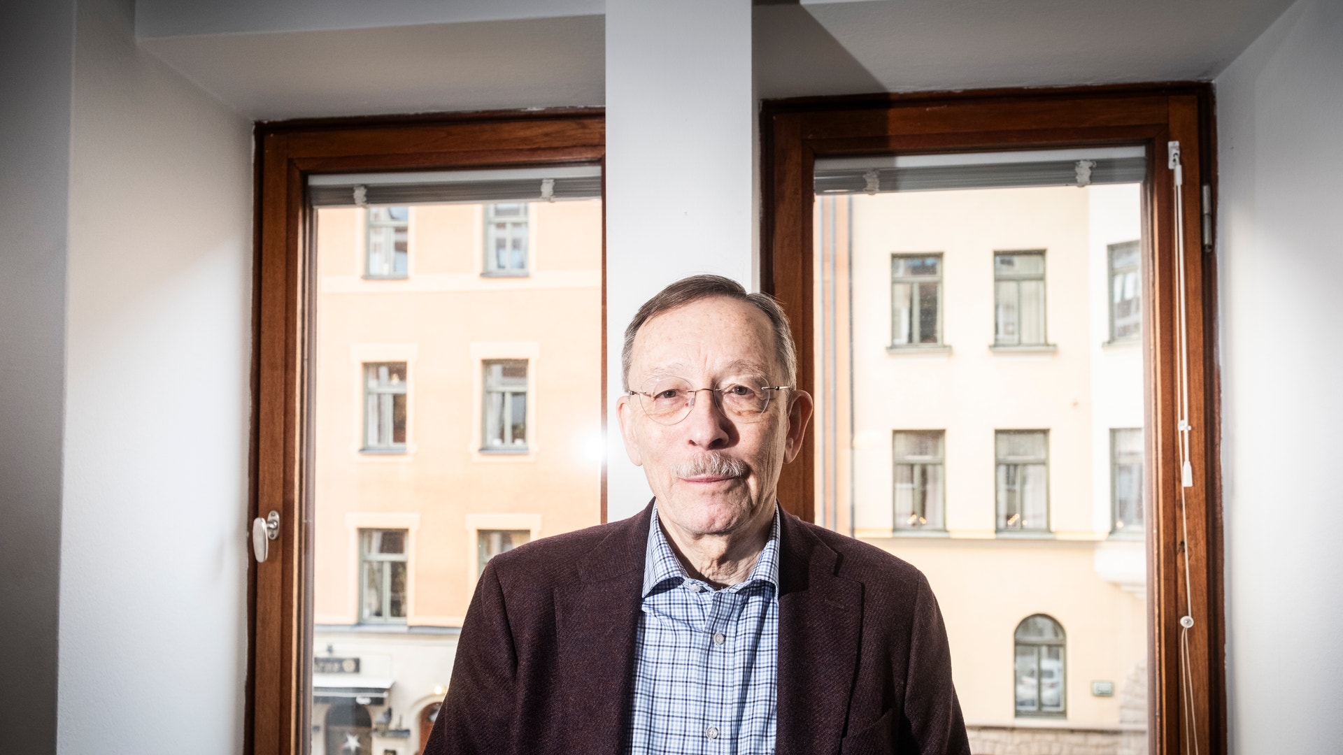 Porträtt av en äldre man med glasögon och mörkbrun kavaj som står framför ett fönster med utsikt över en stadsbyggnad.