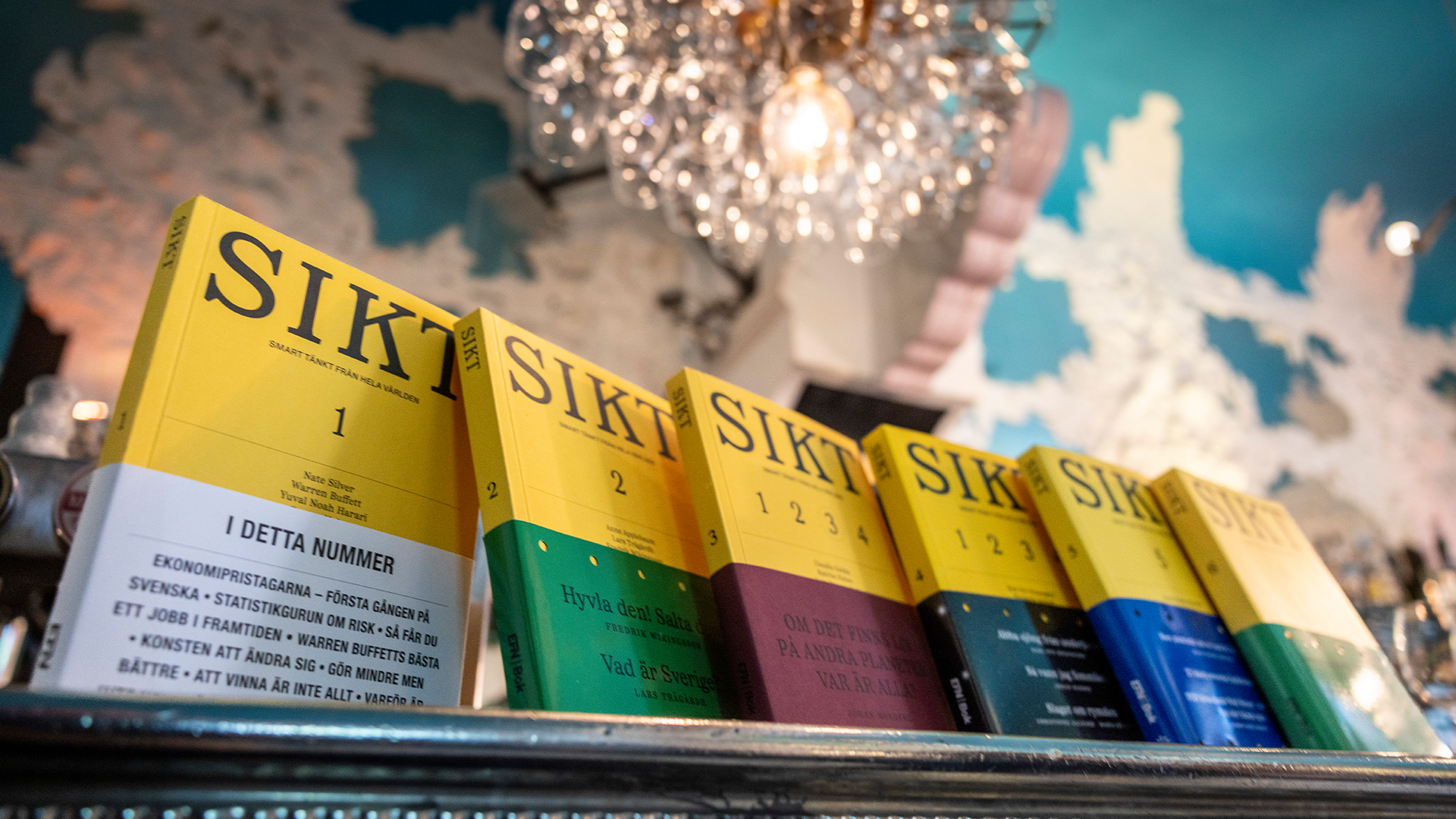 En rad gula böcker märkta SIKT på en hylla, varje bok med en färgad nedre del i olika färger. En dekorativ glaskula syns i bakgrunden mot en blå vägg.