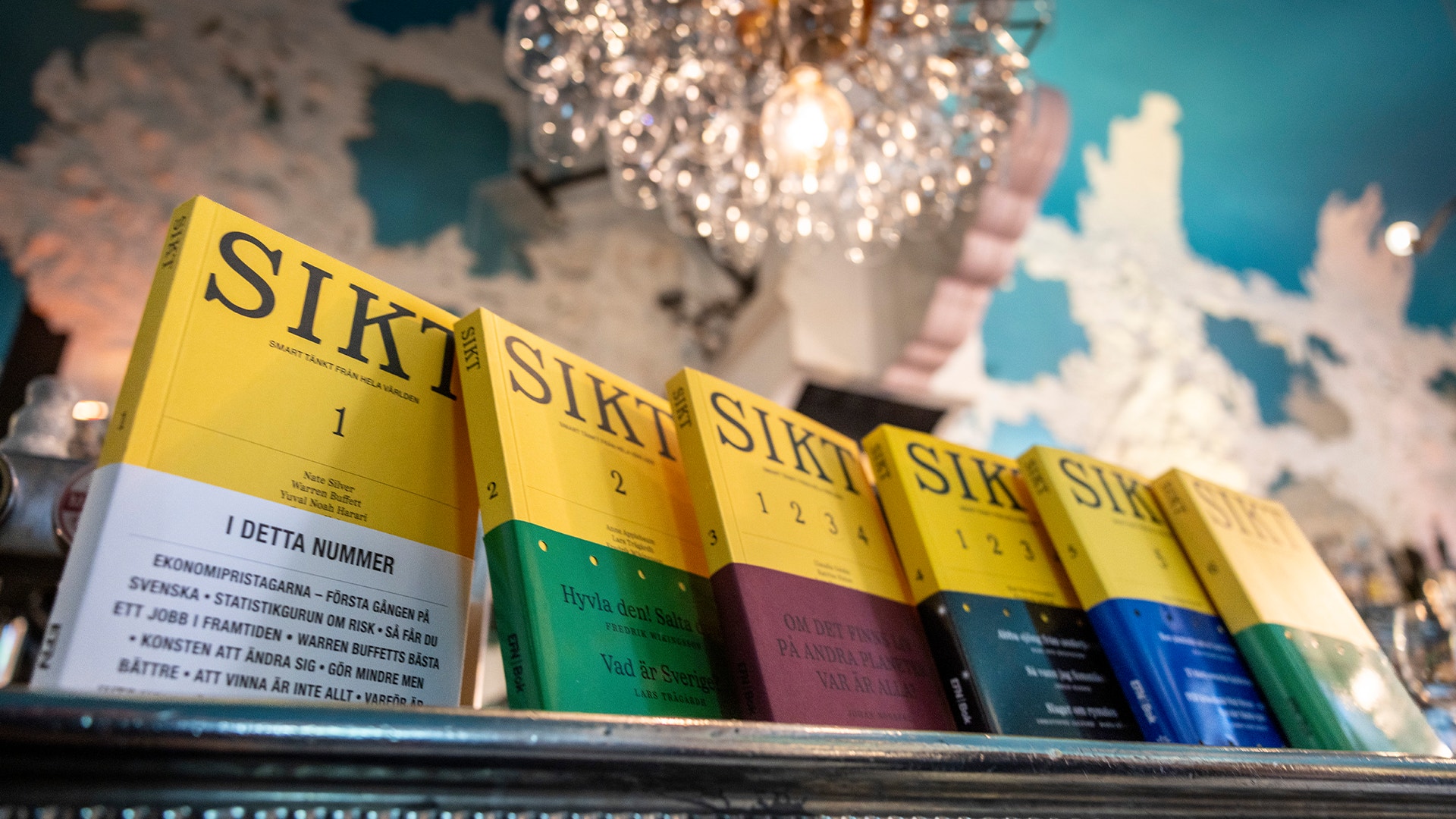 En rad gula böcker märkta SIKT på en hylla, varje bok med en färgad nedre del i olika färger. En dekorativ glaskula syns i bakgrunden mot en blå vägg.