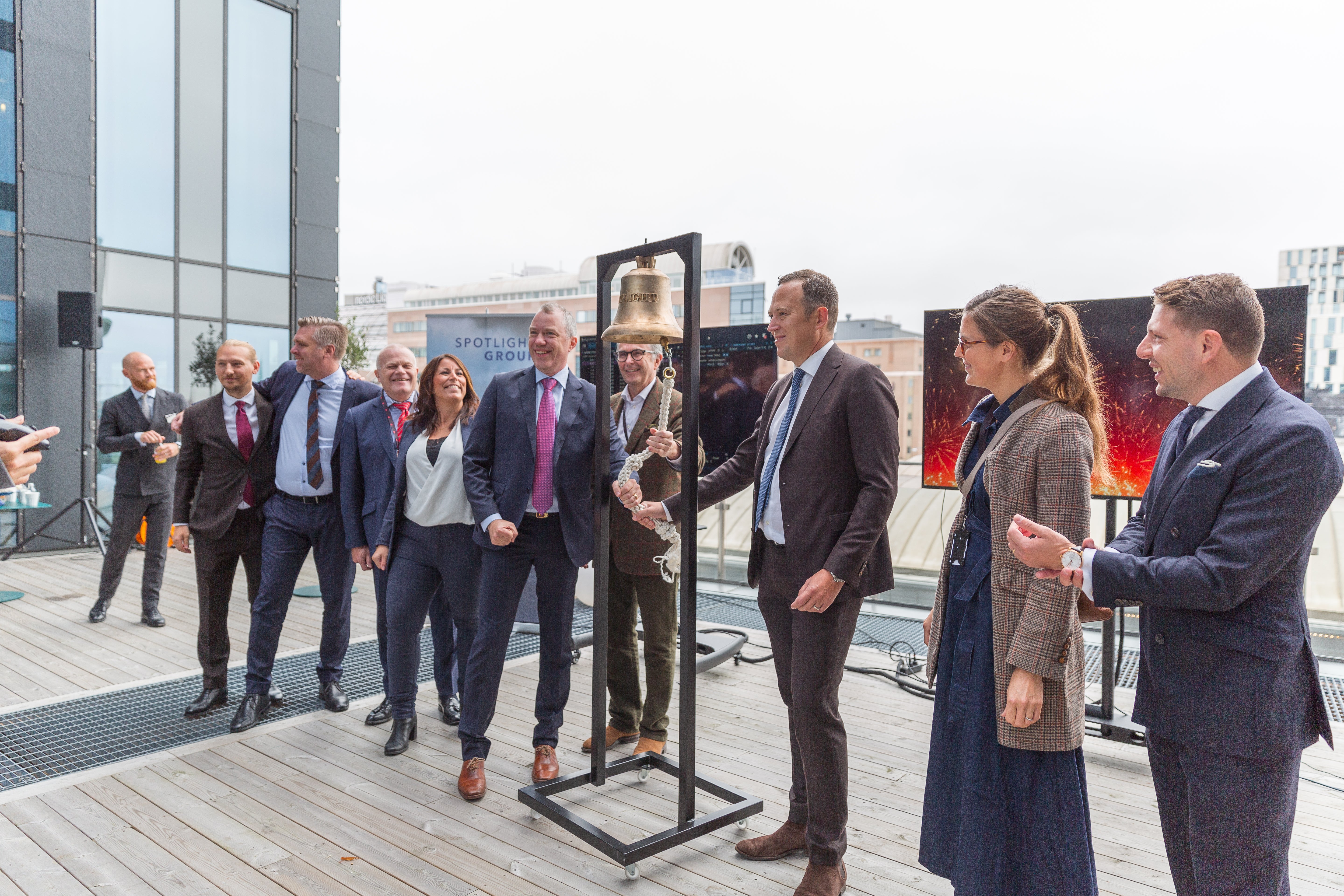 En grupp människor i formella kläder står på en utomhusterrass med stadsvy i bakgrunden. De verkar delta i ett officiellt event eller möte.