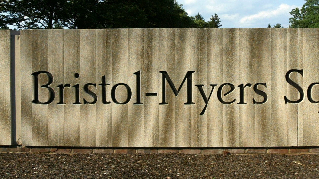 Skylt för läkemedelsföretaget Bristol-Myers Squibb med företagslogotyp och namn på ljusgrå bakgrund, omgiven av gröna träd.
