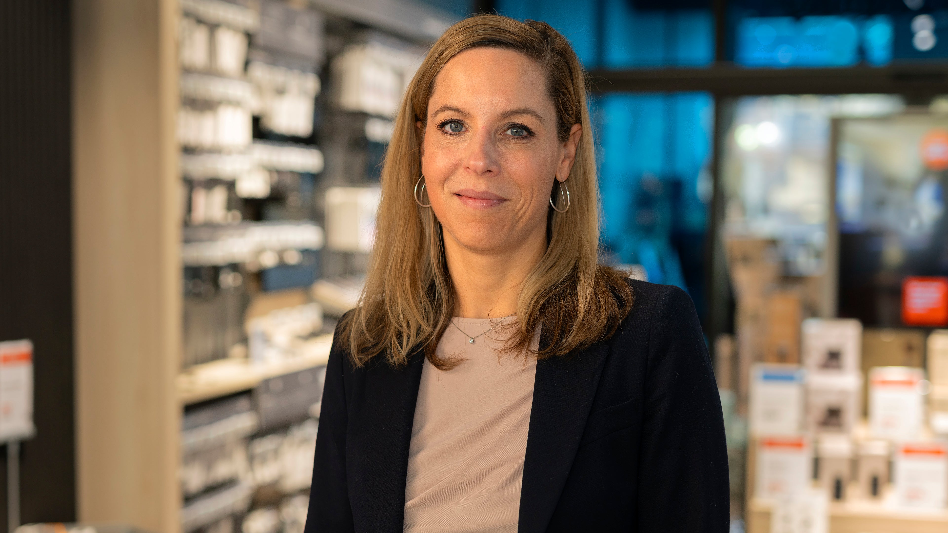 Porträtt av en kvinna i professionell miljö med moderna butikshyllor i bakgrunden.