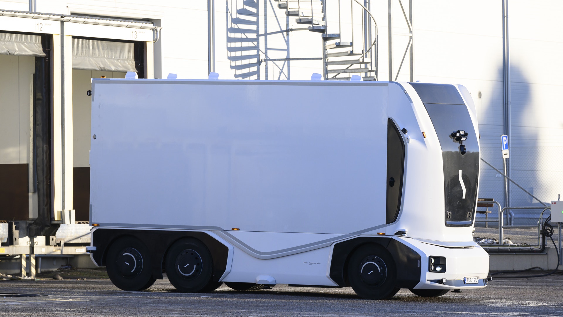 En blå och vit autonom leveransrobot med sex hjul parkerad vid en industribyggnad.