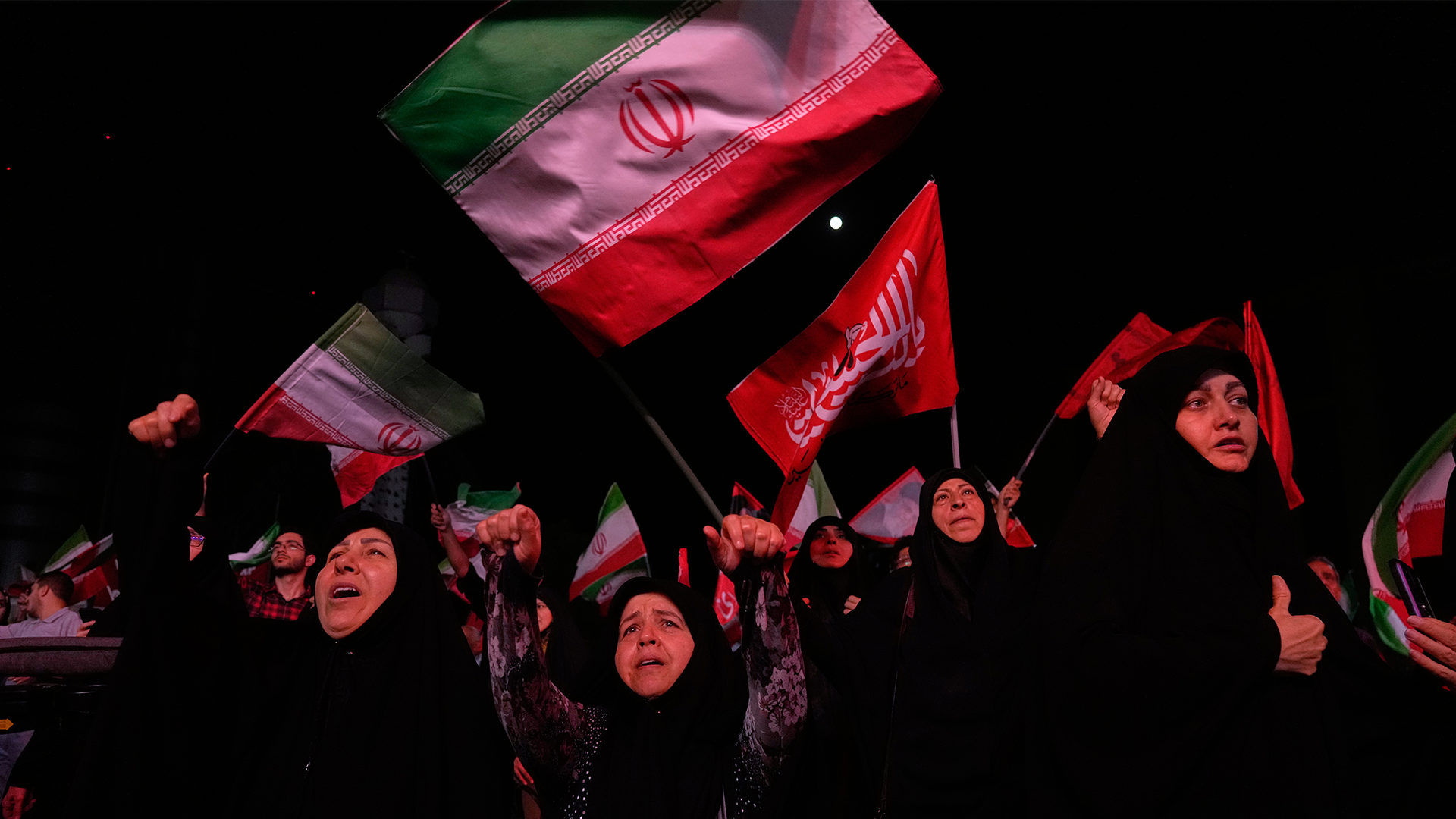 Folkmassa som håller upp Irans flagga och röda flaggor under en demonstration mot mörk bakgrund.