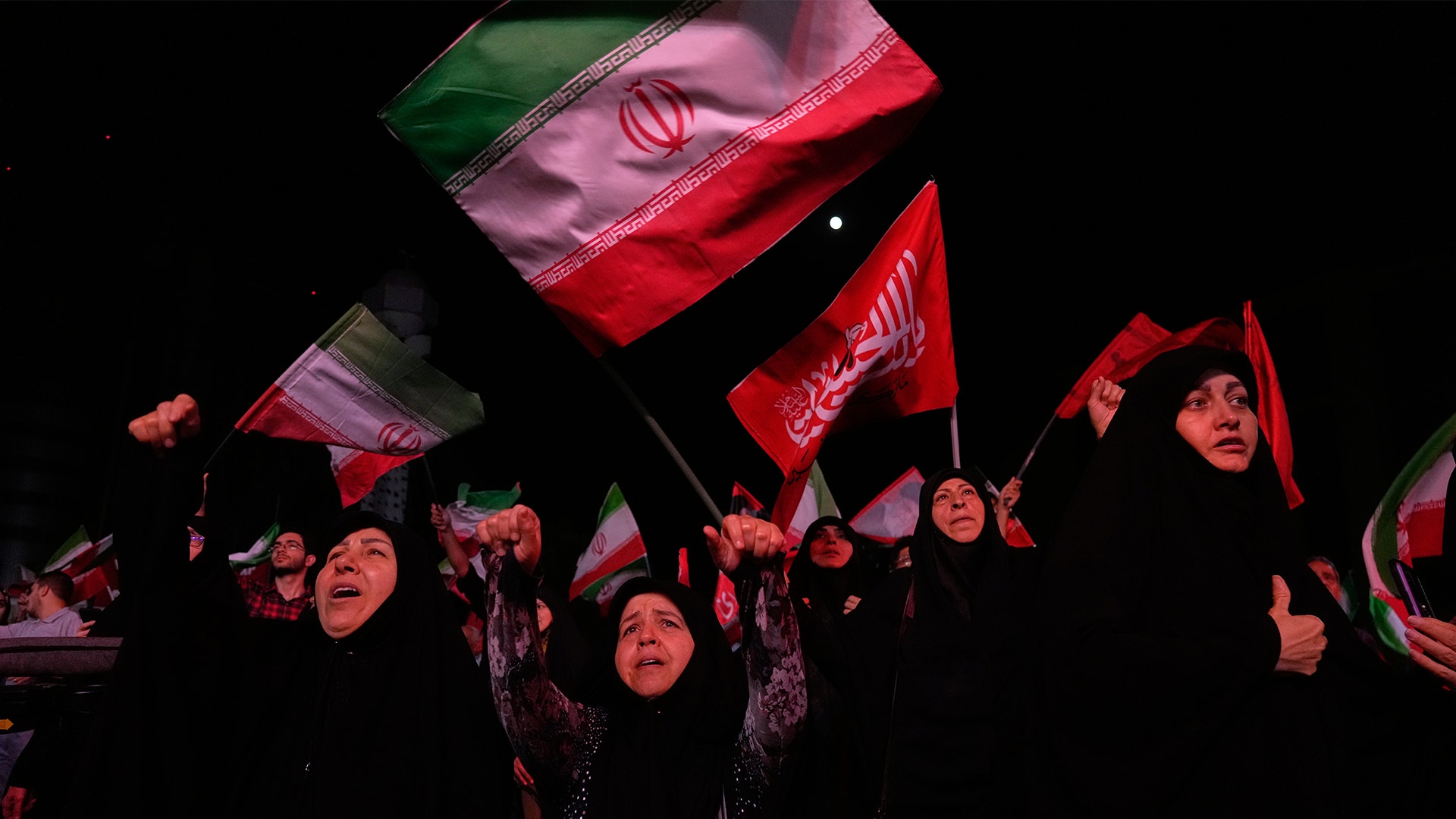 Folkmassa som håller upp Irans flagga och röda flaggor under en demonstration mot mörk bakgrund.