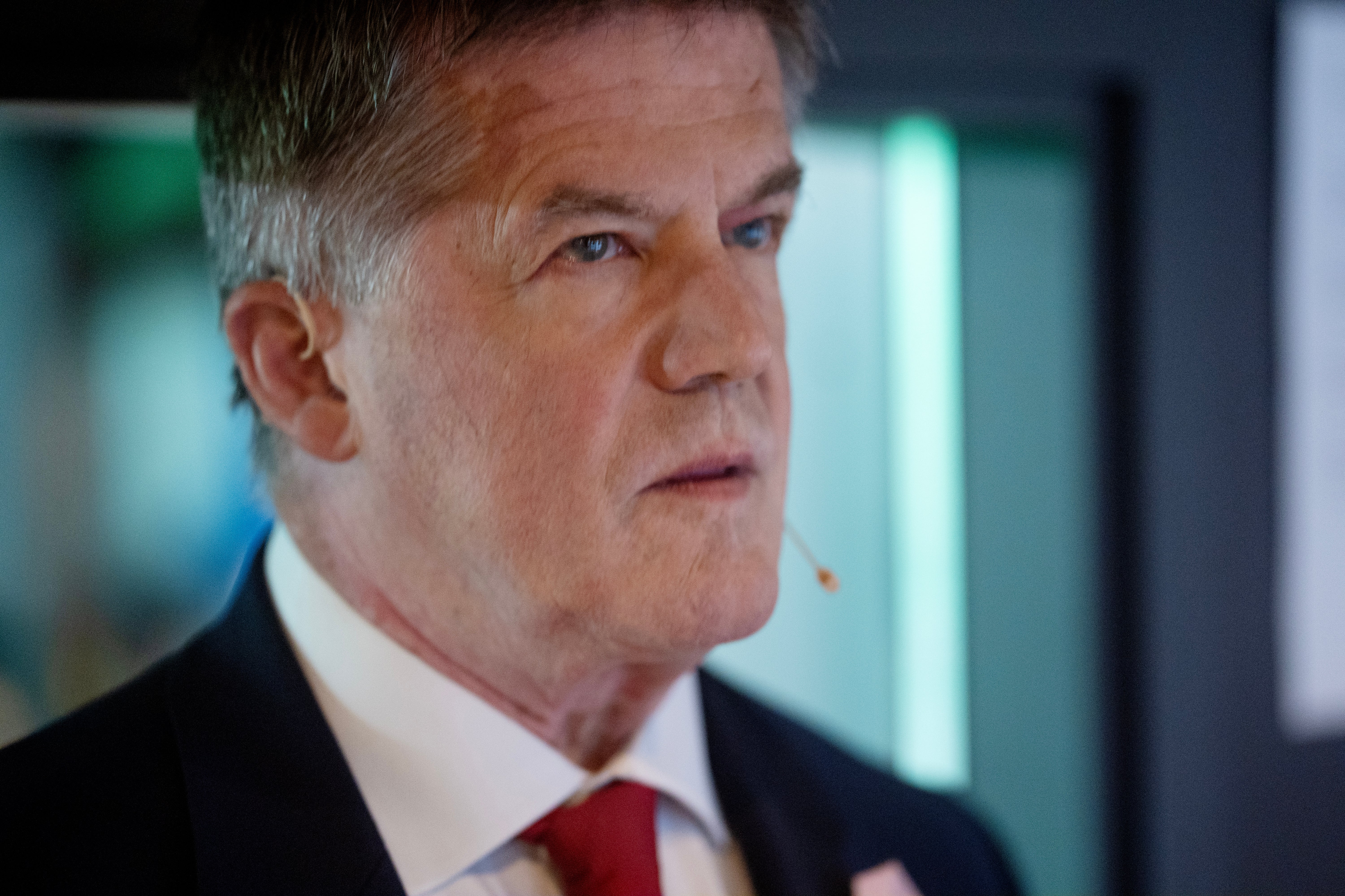 Närbild på Ilja Batljan, en äldre man i mörk kostym, vit skjorta och röd slips, med en liten mikrofon vid kinden.