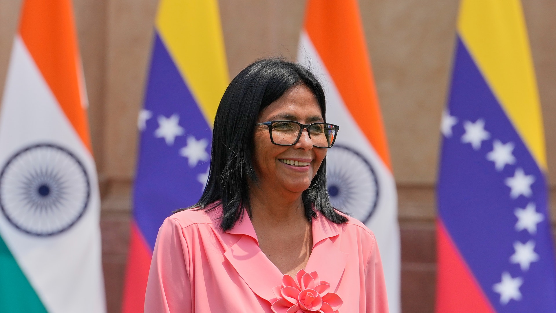 Kvinna i rosa skjorta med blomsterbrosch framför Indiens och Venezuelas flaggor.
