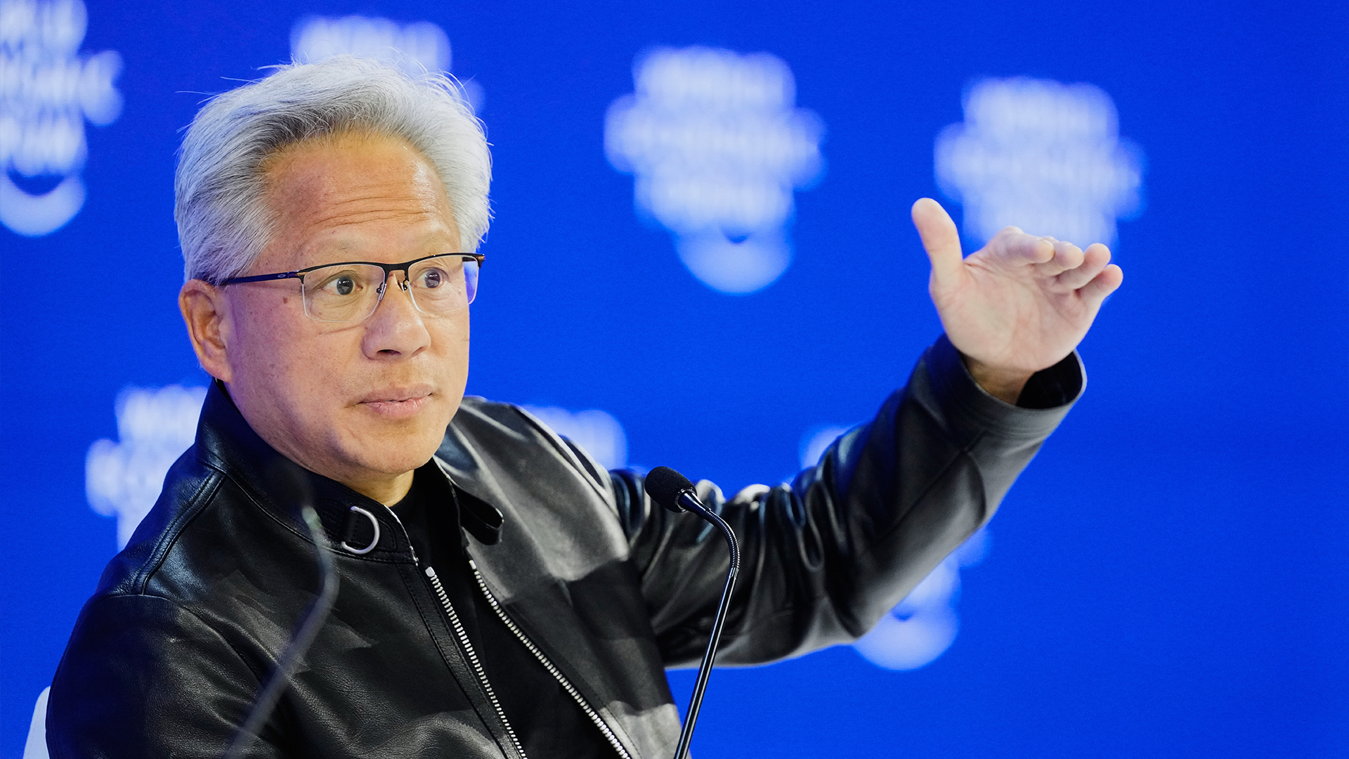 Man i svart läderiacka som gestikulerar under en presentation mot blå bakgrund