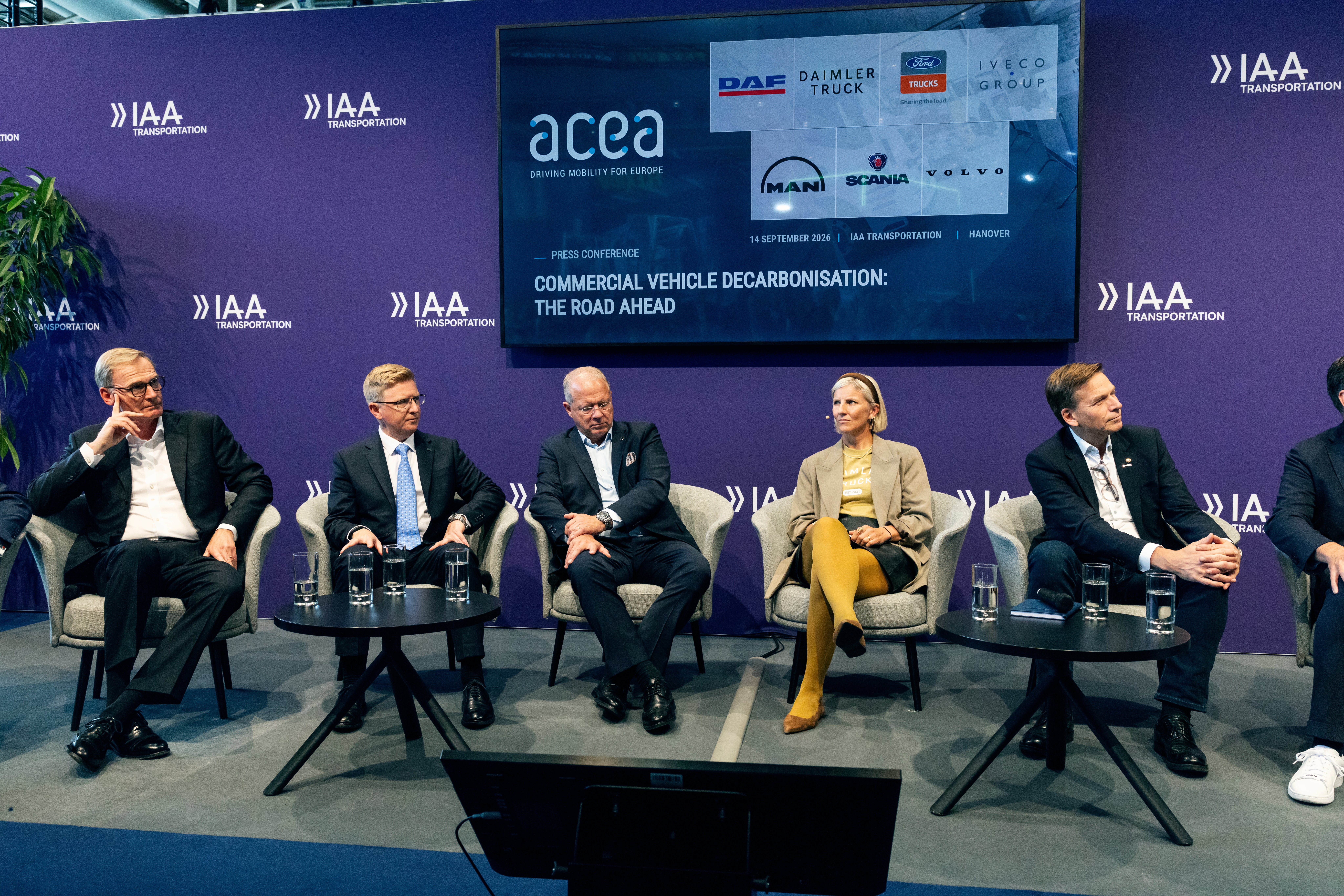 Fem personer i kostym sitter i fåtöljer på en scen under en pressträff, med en skärm bakom sig som visar rubriken "Commercial vehicle decarbonisation: the road ahead" och logotyper från fordonstillverkare.