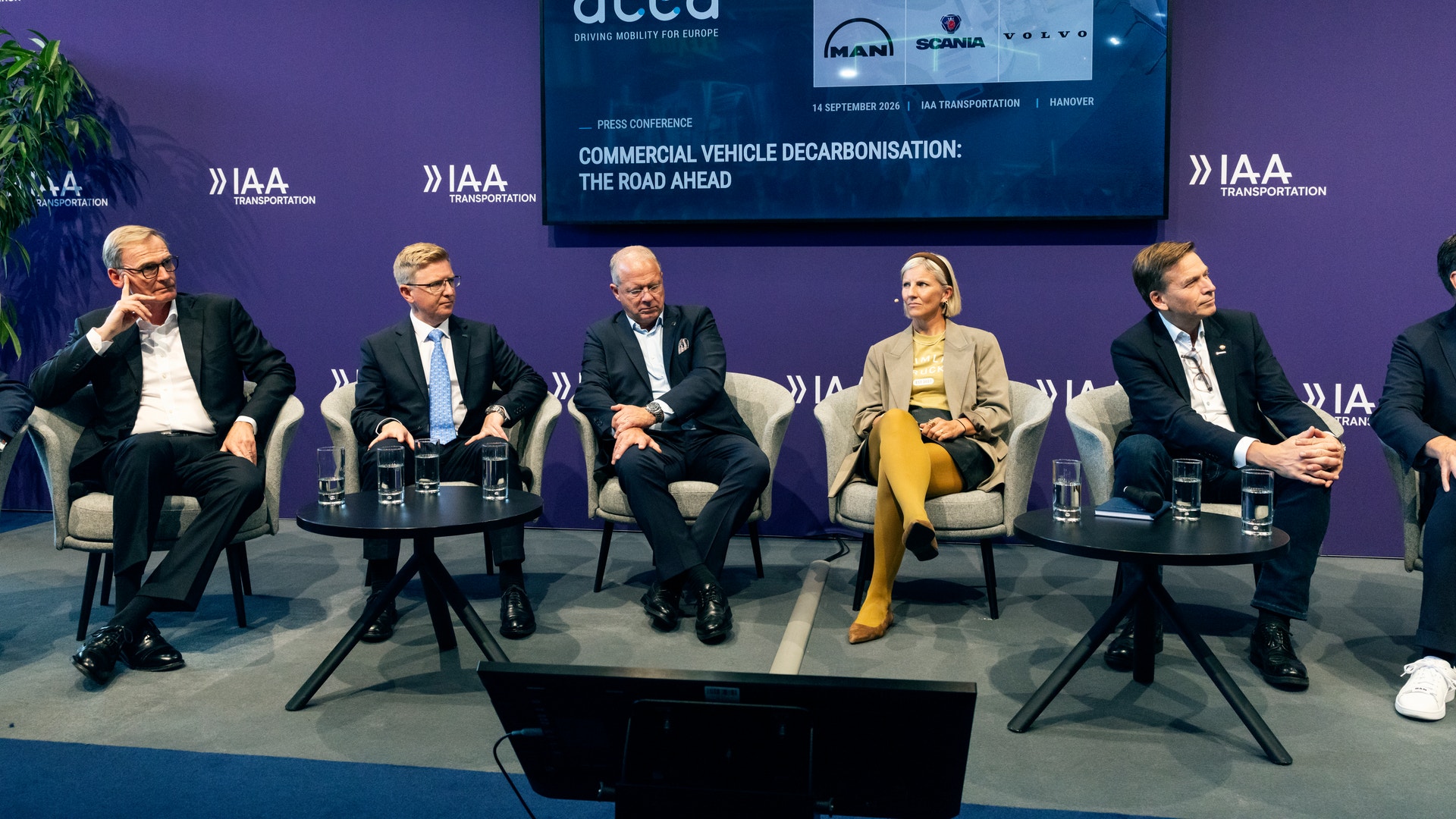 Fem personer i kostym sitter i fåtöljer på en scen under en pressträff, med en skärm bakom sig som visar rubriken "Commercial vehicle decarbonisation: the road ahead" och logotyper från fordonstillverkare.