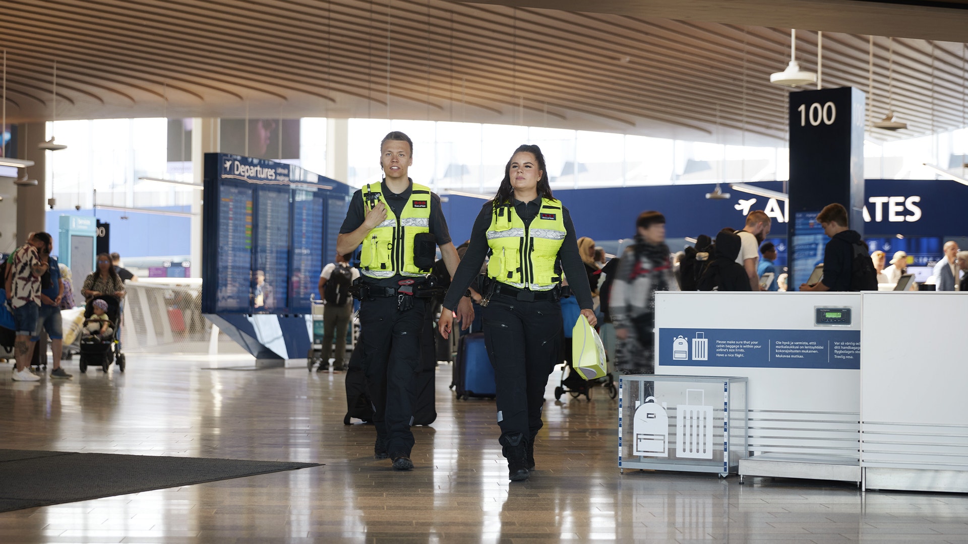 Två väktare i reflexvästar går genom en flygplatsterminal med resenärer i bakgrunden.