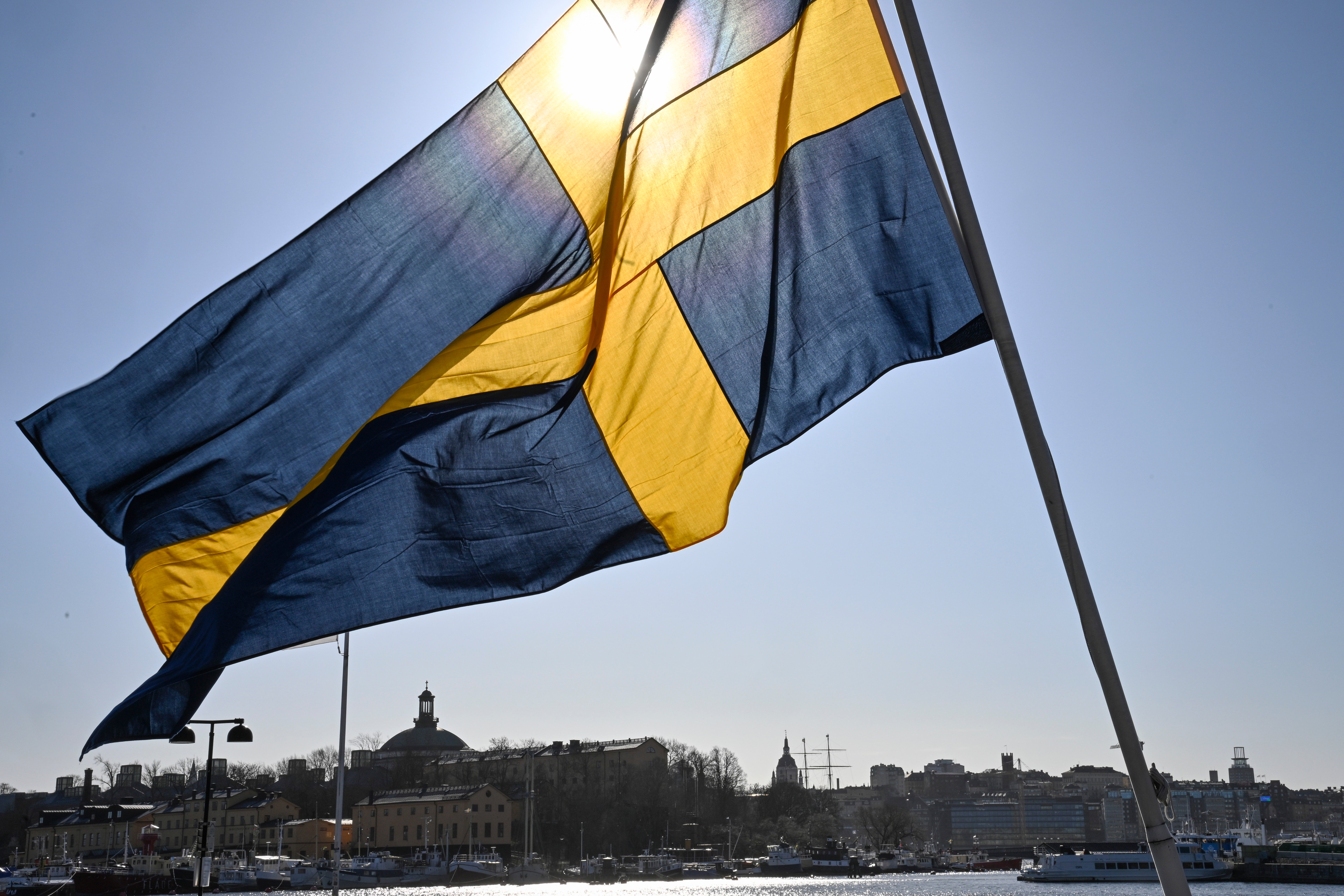 Sveriges flagga med blått och gult viftar i vinden. I bakgrunden syns en hamn med båtar och en stad med byggnader och kyrktorn.
