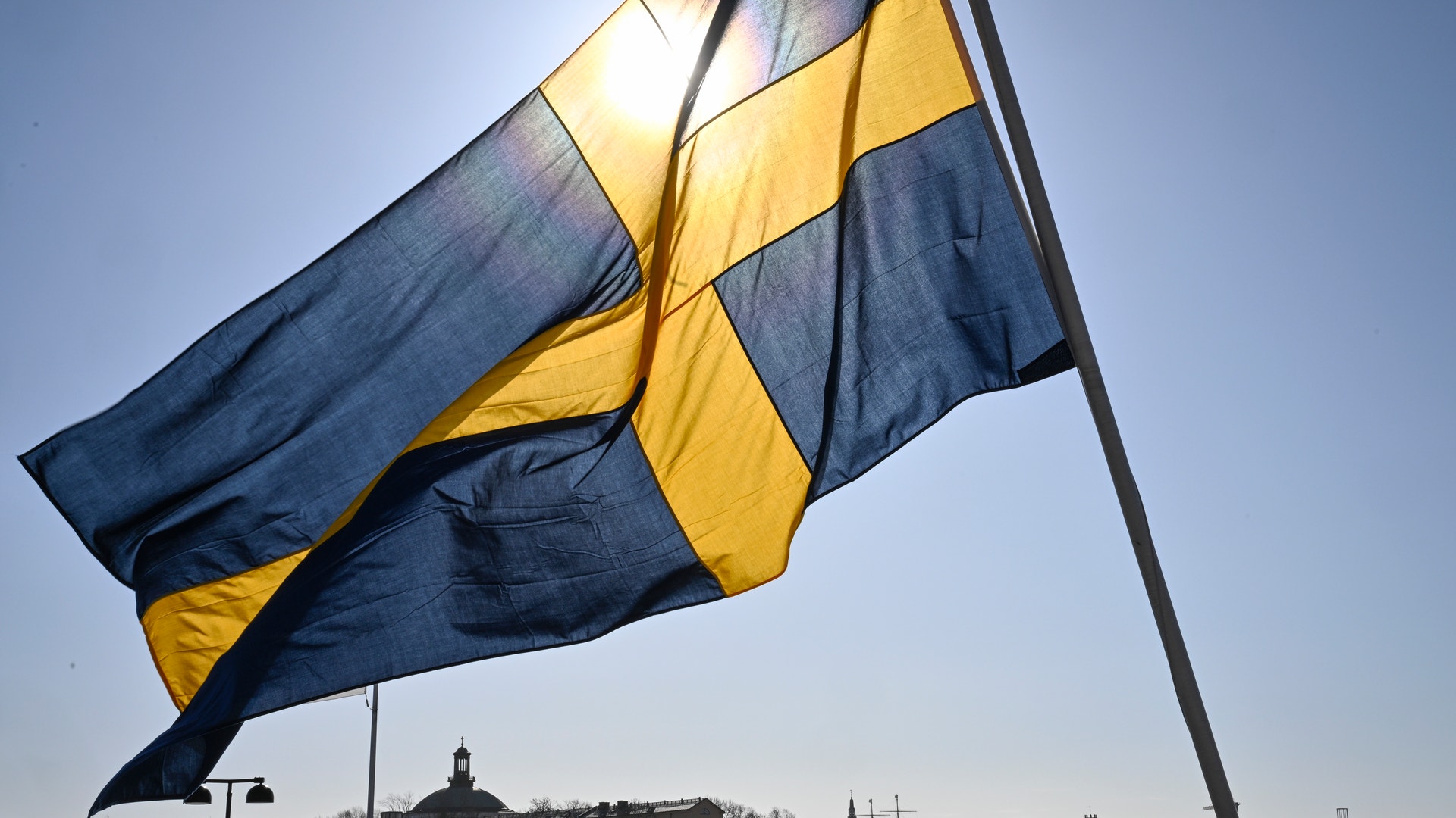 Sveriges flagga med blått och gult viftar i vinden. I bakgrunden syns en hamn med båtar och en stad med byggnader och kyrktorn.
