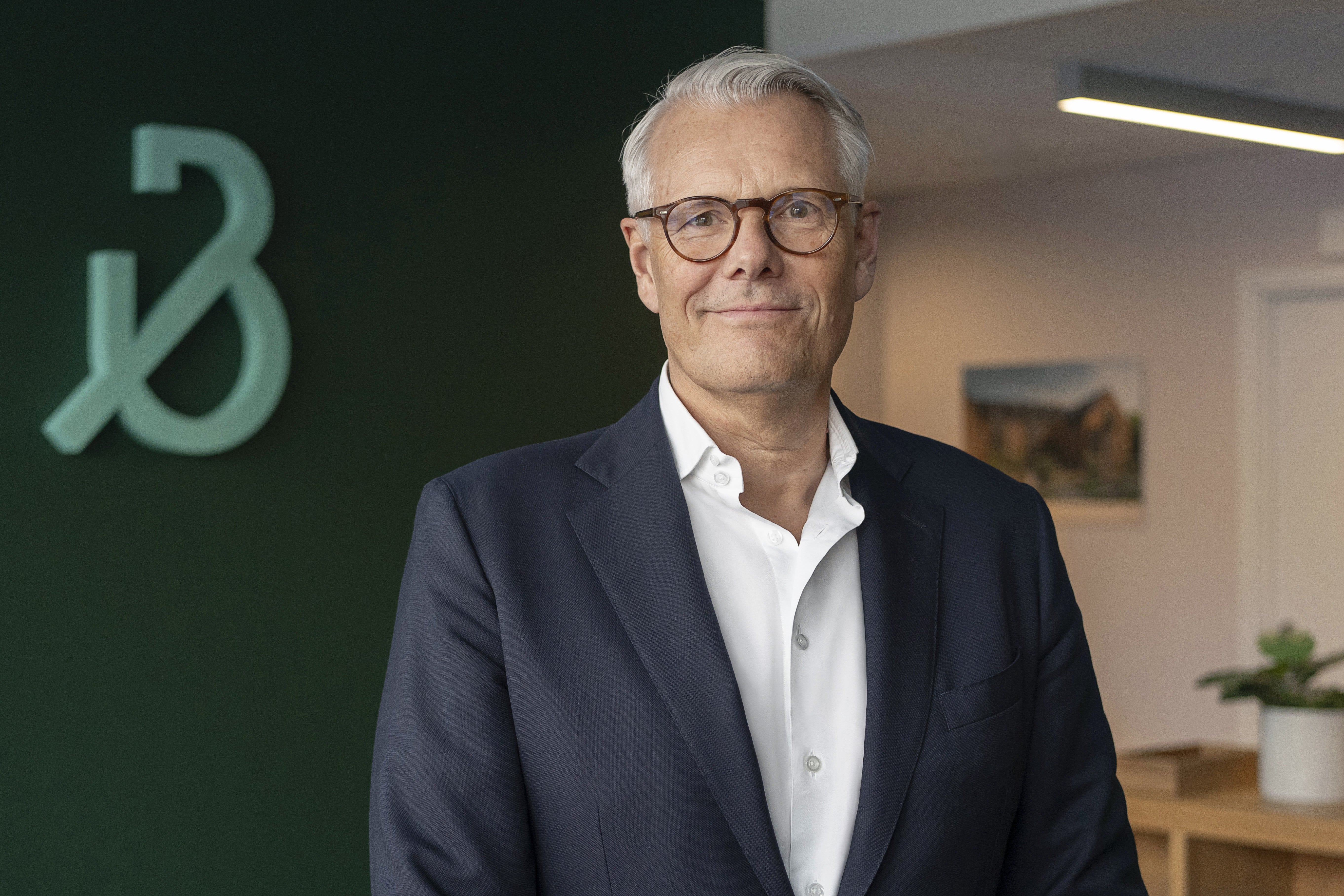 Porträtt av Peter Wallin, verkställande direktör för Bonava. Han är en äldre man med grått hår och glasögon, klädd i mörkblå kavaj och vit skjorta. Han står framför en grön vägg med Bona-logotypen och ler mot kameran.