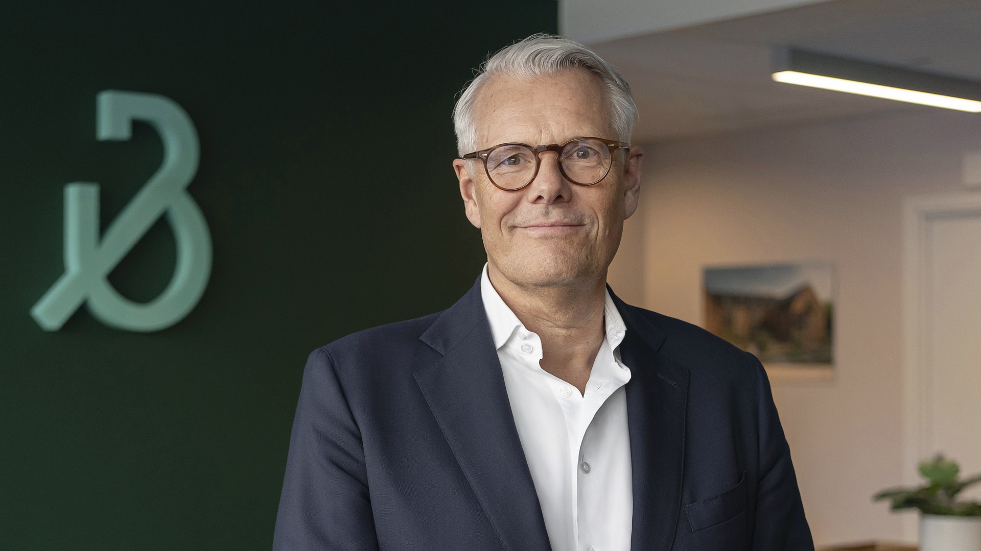 Porträtt av Peter Wallin, verkställande direktör för Bonava. Han är en äldre man med grått hår och glasögon, klädd i mörkblå kavaj och vit skjorta. Han står framför en grön vägg med Bona-logotypen och ler mot kameran.