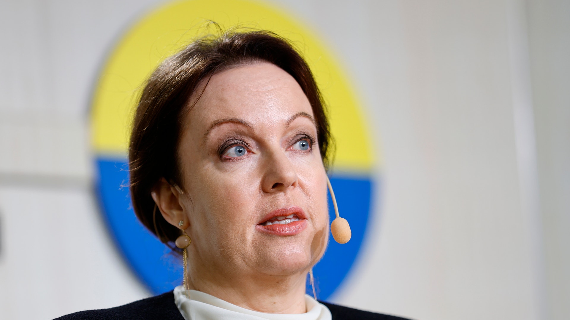 Närbild på en kvinna med mörkt hår och headsetmikrofon som talar, framför en blå och gul bakgrund.