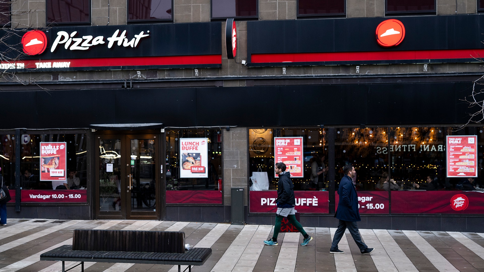Exteriör av en Pizza Hut-restaurang med svart fasad och röd logotyp. Människor går förbi på en stadsgata framför restaurangen.