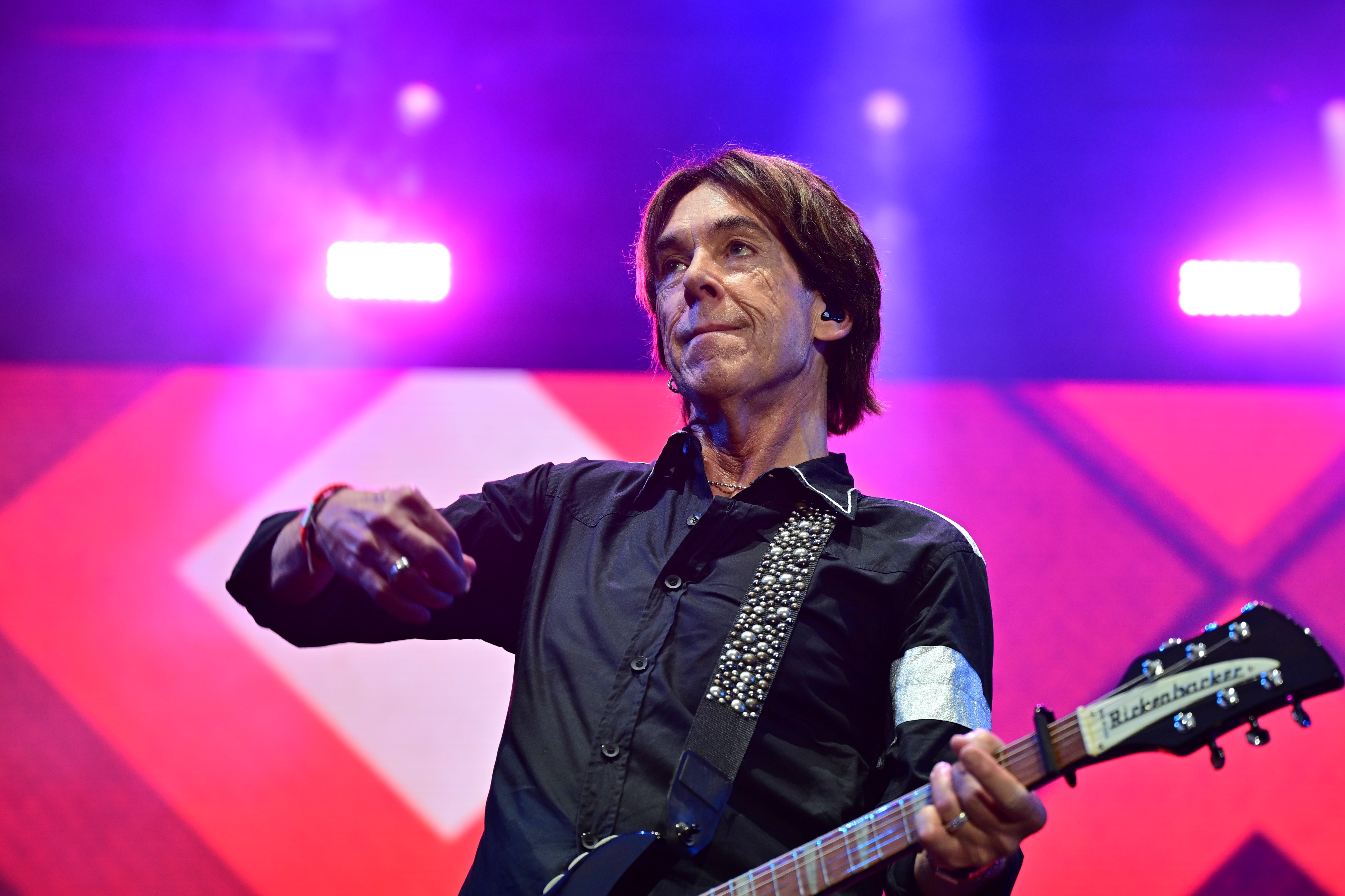 Per Gessle står på en scen med en gitarr och blickar åt sidan, framför en färgglad rosa och lila bakgrund.